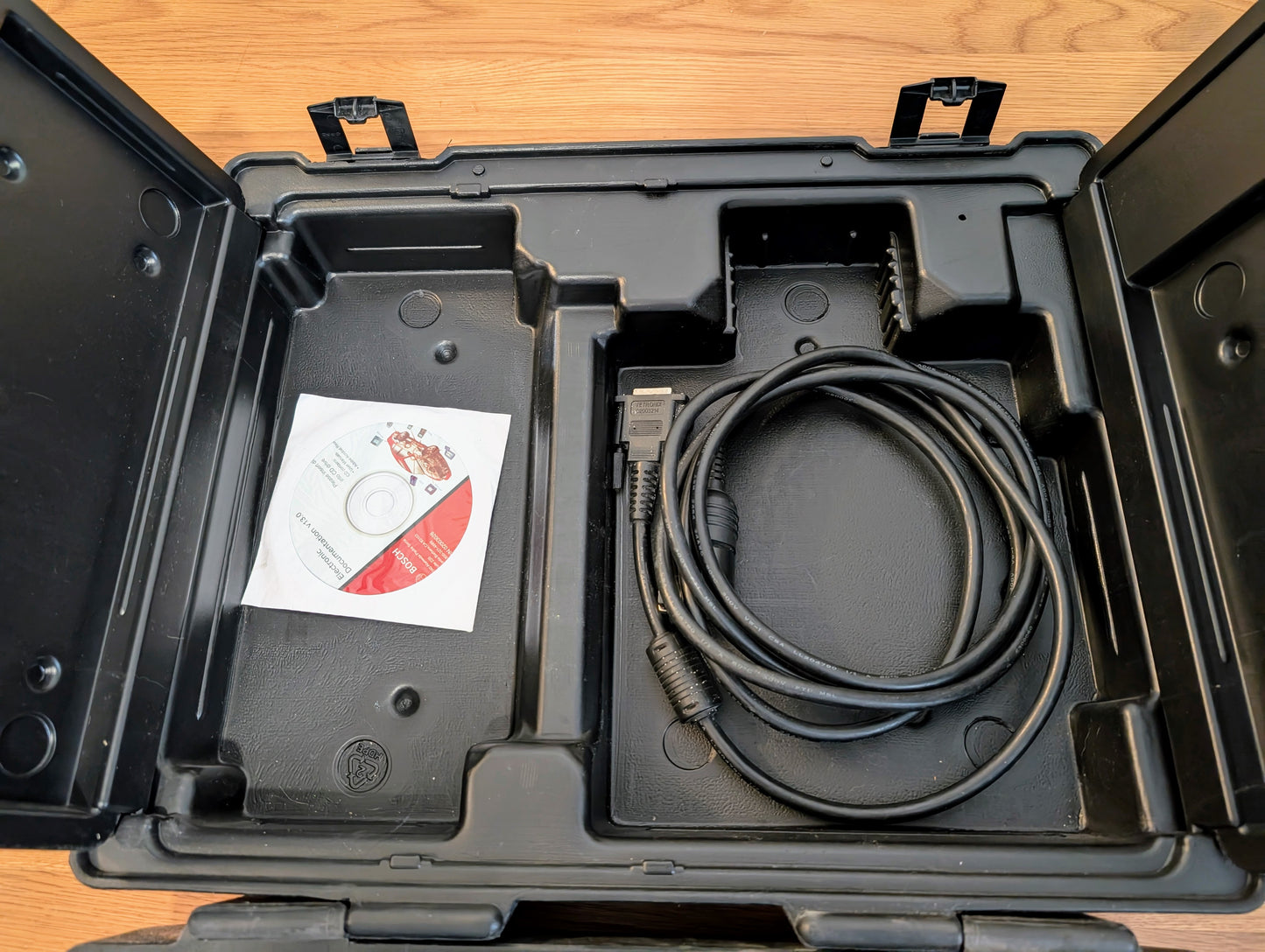 An open black Vetronix case on a wooden surface holds a coiled cable with connectors and a CD, designed for use with the TESTED Tech 2 Automotive Diagnostic Tool & Accessories for 1991-2013 GM vehicles. The case has organizing compartments.