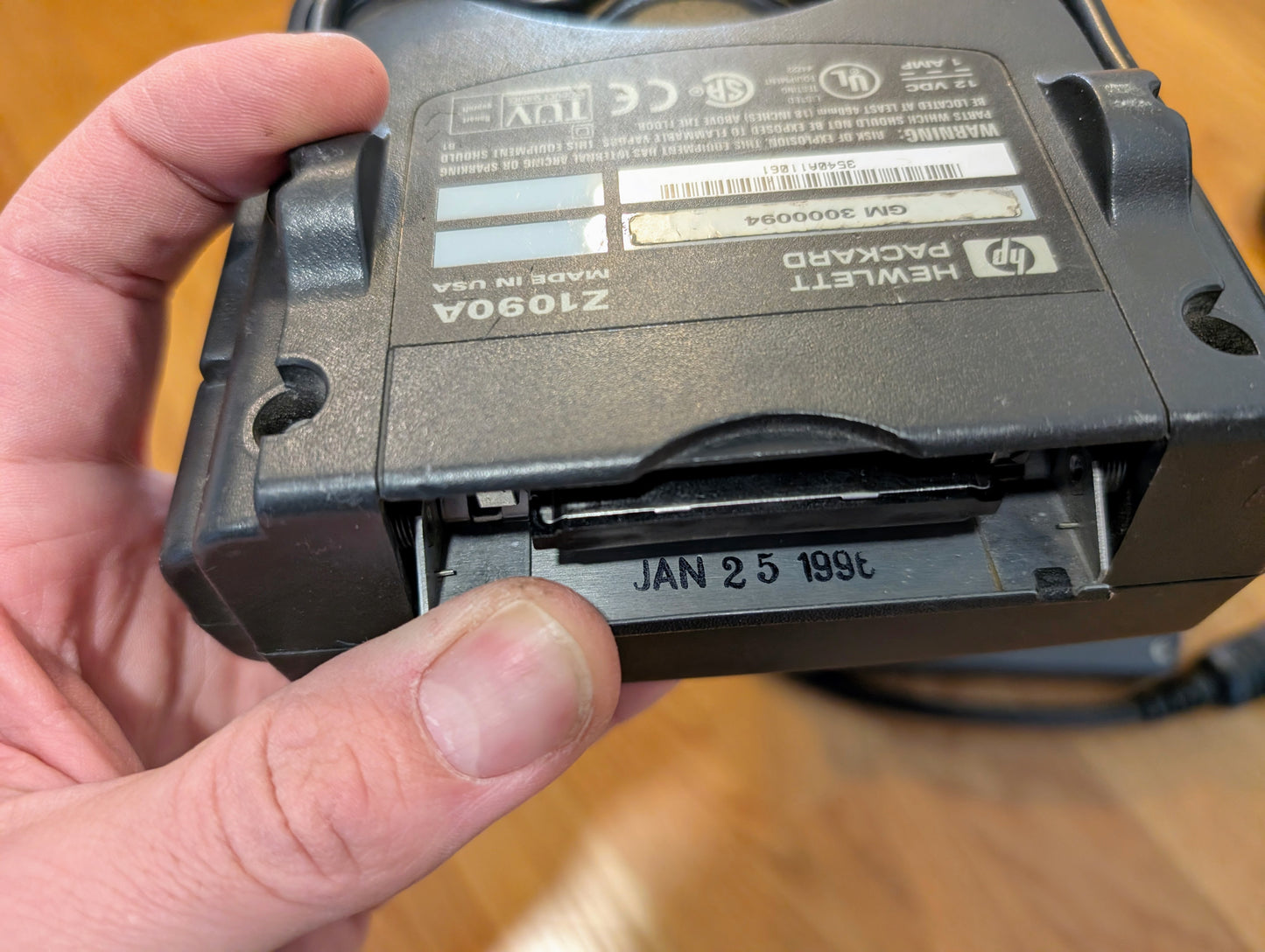 A close-up of a hand holding a Vetronix TESTED Tech 2 Automotive Diagnostic Tool & Accessories for 1991-2013 GM Vehicles, showing a visible date code JAN 25 1996 inside a compartment and product labels on the back, resting on wood.