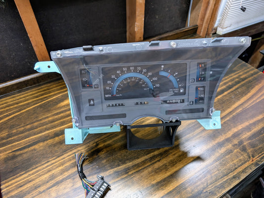 A General Motors OEM Instrument Cluster with PRNDL and 110 mph speedo for 1986-1994 Chevy S10 Blazer rests on a wooden table, partially covered by a clear protective sheet, with the wiring harness visible in the foreground.
