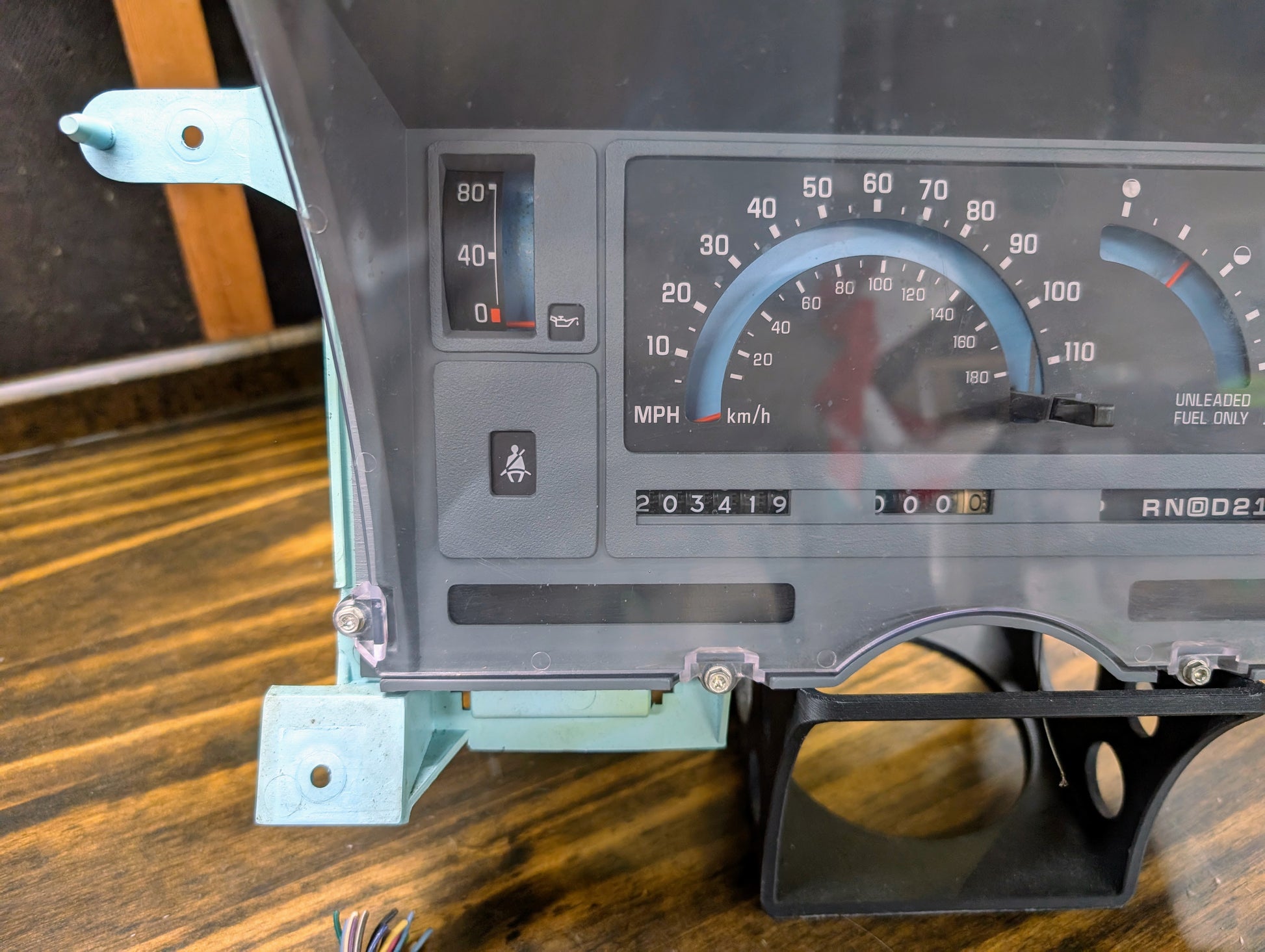 Close-up of a General Motors OEM Instrument Cluster with PRNDL and 110 mph speedo for 1986-1994 Chevy S10 Blazer, showing 203419 miles on the odometer, illuminated oil pressure and seatbelt lights, displayed on a wooden surface.