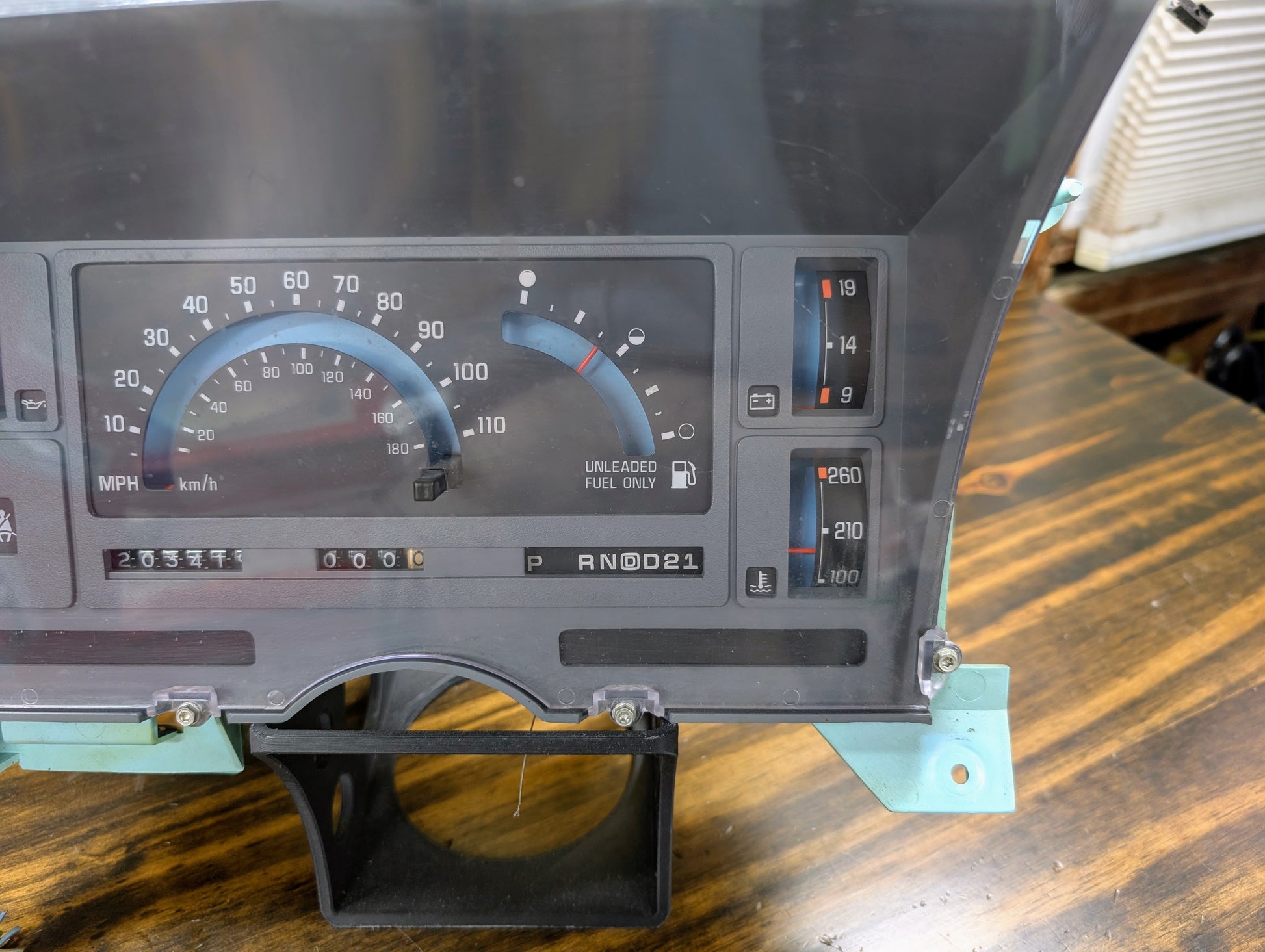 Close-up of a General Motors OEM Instrument Cluster with PRNDL and 110 mph speedo for 1986-1994 Chevy S10 Blazer, showing speedometer, fuel and temp gauges, odometer at 203587 miles, and gear indicator on a wooden surface.