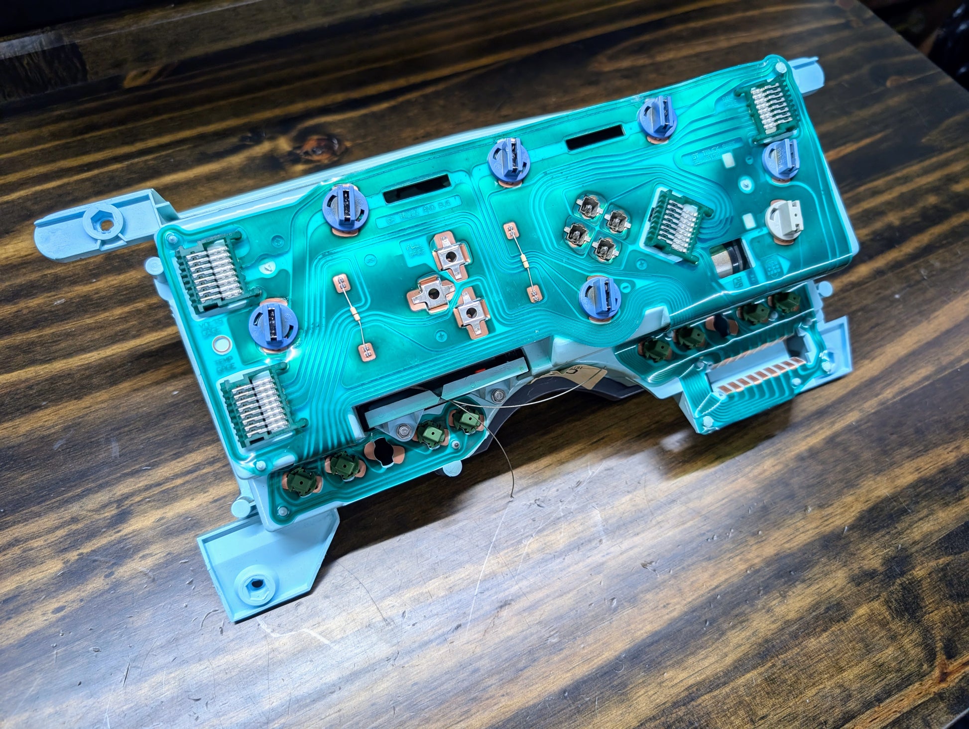 A close-up of a disassembled General Motors OEM Instrument Cluster with PRNDL and 110 mph Speedo for 1986-1994 Chevy S10 Blazer, showing its green printed circuit board, copper traces, and connectors on a wooden surface.