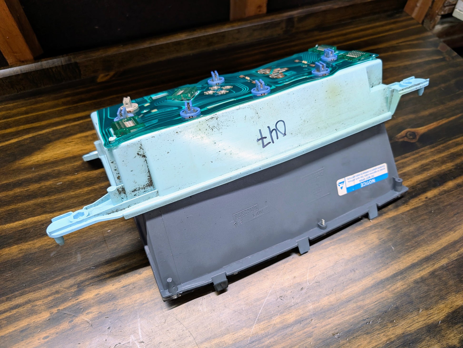 A General Motors OEM Instrument Cluster with PRNDL and 110 mph Speedo for 1986-1994 Chevy S10 Blazer is shown on a wooden surface, displaying its casing, internal circuit board, signs of use, and a handwritten side marking.