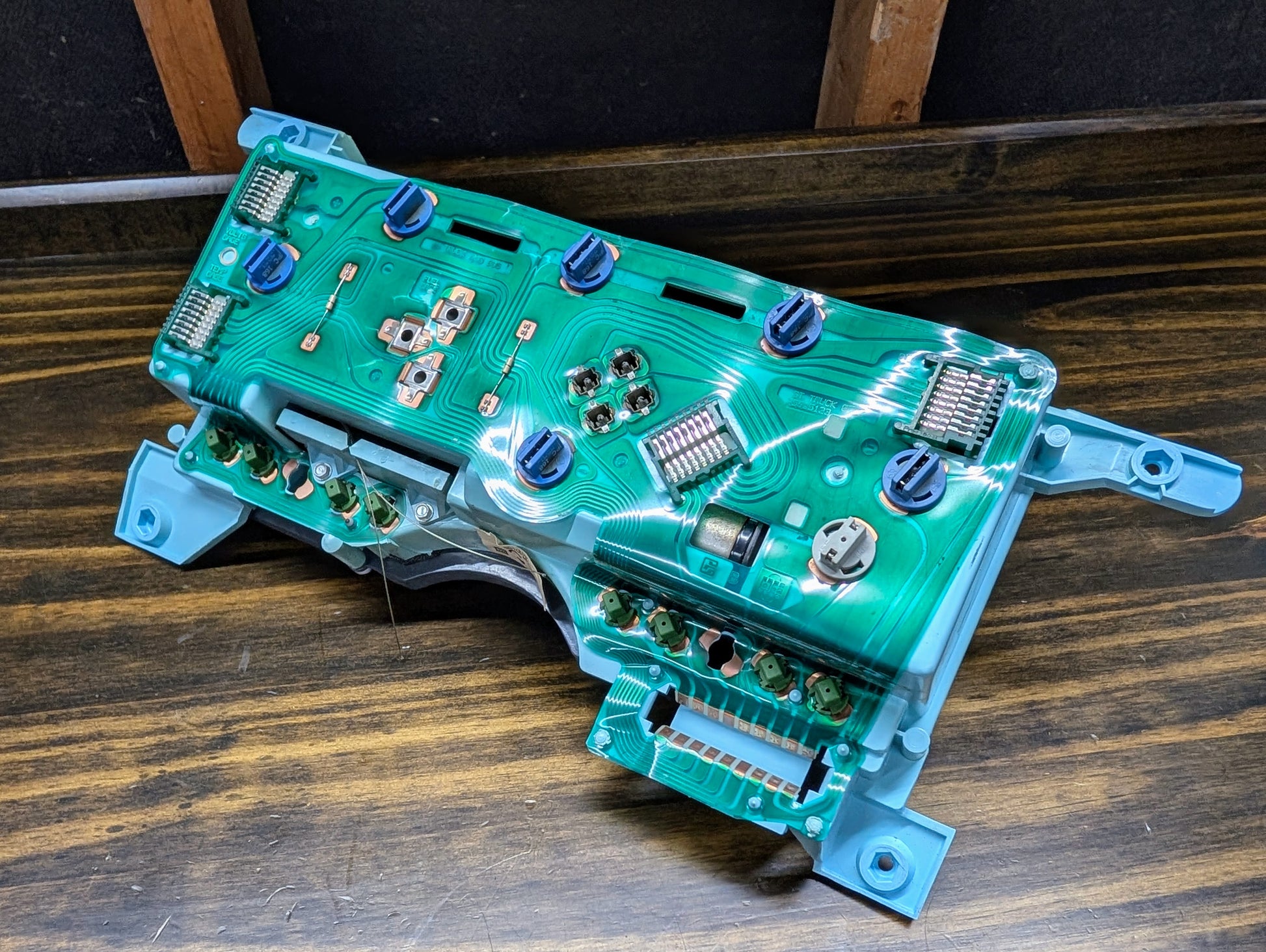 A close-up of a General Motors OEM Instrument Cluster with PRNDL and 110 mph Speedo for 1986-1994 Chevy S10 Blazer, showing green circuitry and blue connectors on a wooden workbench with dark walls in the background.