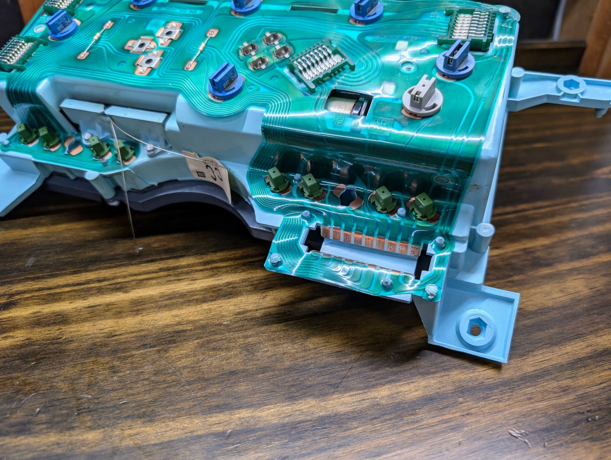 Close-up of a General Motors OEM Instrument Cluster circuit board with green traces, blue connectors, and electronic components for 1986-1994 Chevy S10 Blazer, featuring PRNDL and 110 mph speedo, on a wooden surface.