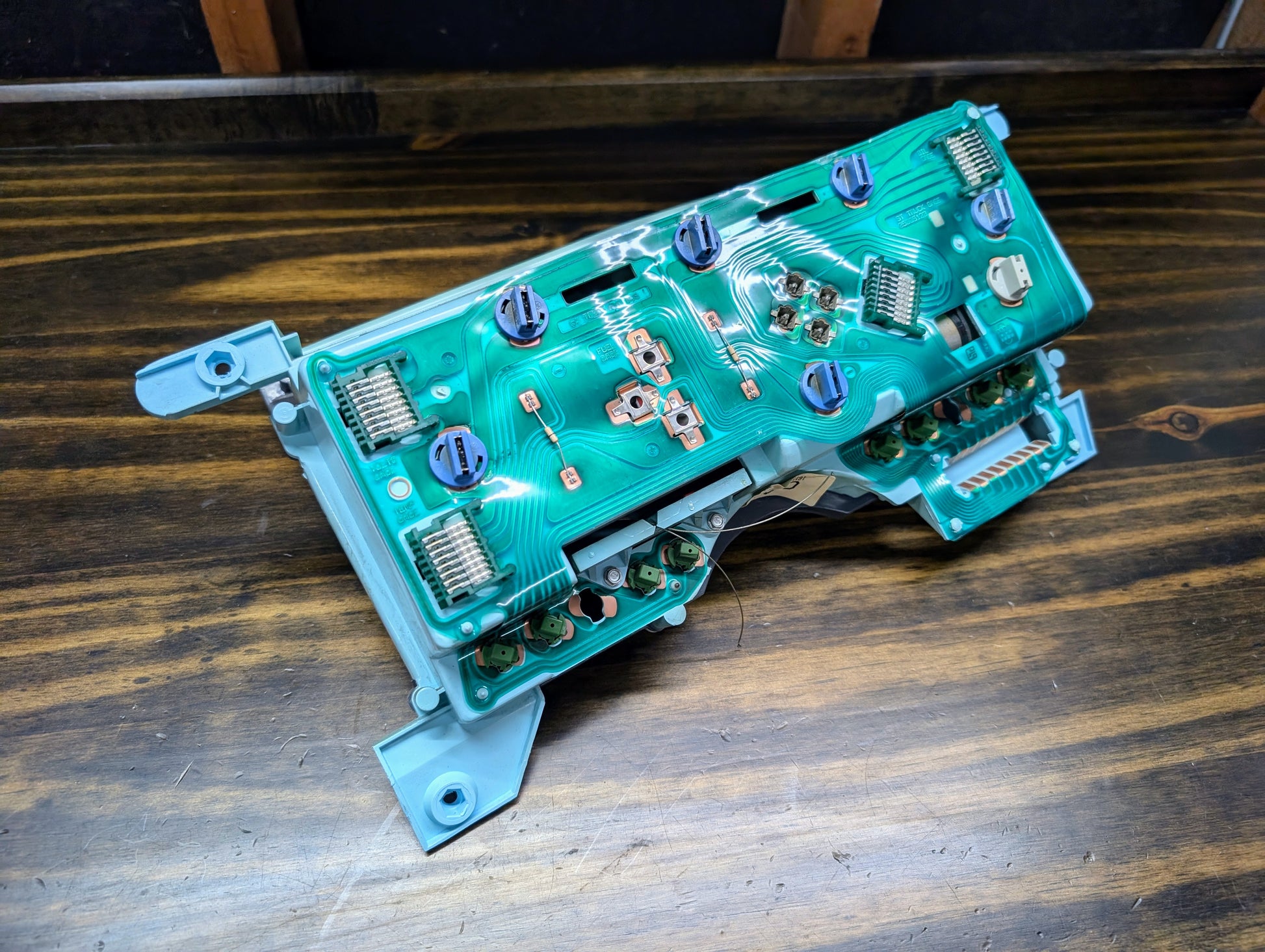 A close-up of a General Motors OEM Instrument Cluster with PRNDL and 110 mph speedo for 1986-1994 Chevy S10 Blazer, showing its green PCB and connectors on a blue frame, resting on wood.