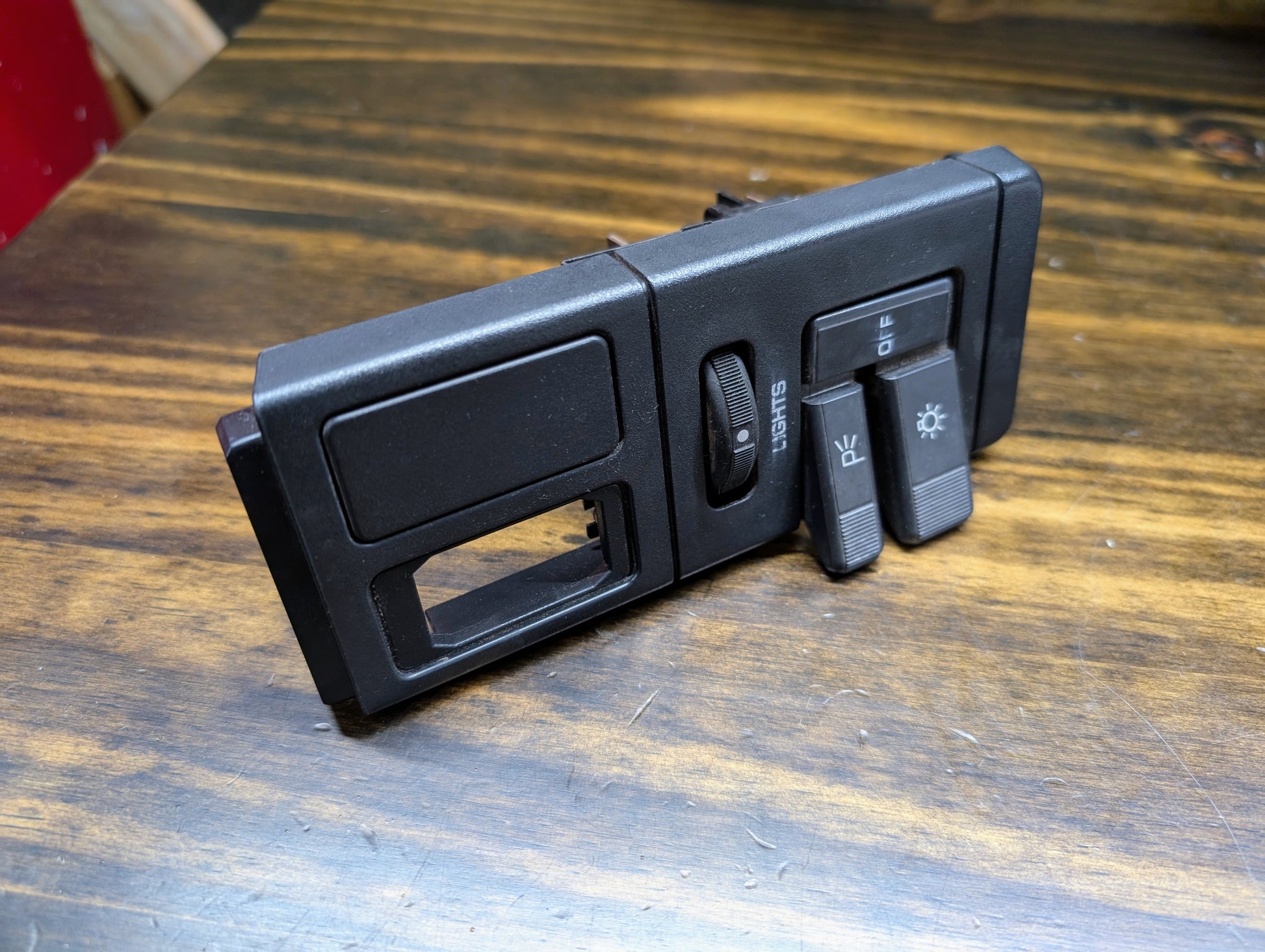 A General Motors OEM Headlight Headlamp Switch in black for 1986-1994 Chevy S10, Blazer, GMC S15, and Jimmy sits on a wooden surface, featuring dimmer and sliding switches with light/fog icons and an empty rectangular slot.