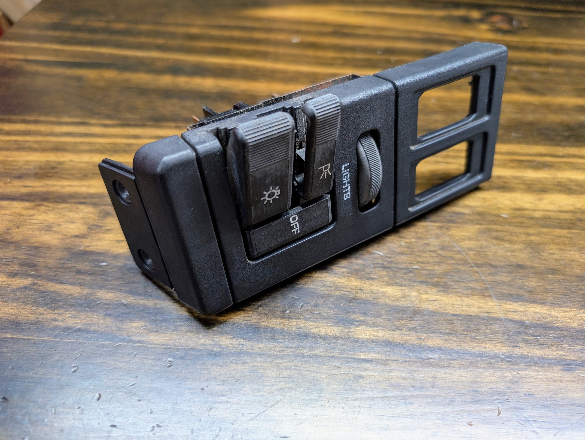 A General Motors OEM Headlight Headlamp Switch in black, designed for 1986–1994 Chevy S10, Blazer, GMC S15, and Jimmy, rests on a worn wooden surface showing slight damage.