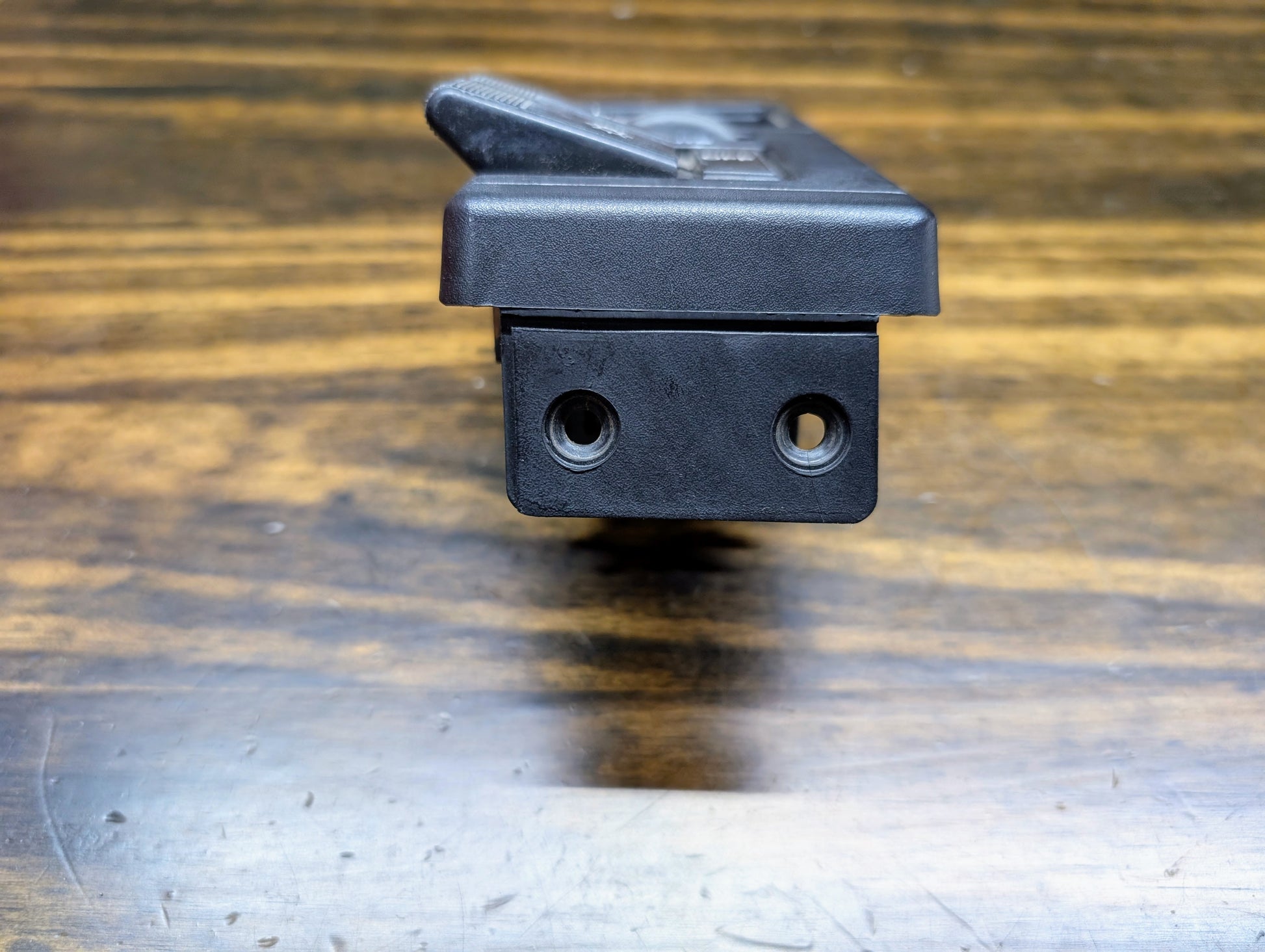 Close-up of a black General Motors OEM Headlight Headlamp Switch for 1986-1994 Chevy S10/Blazer or GMC S15/Jimmy, showing two circular holes at one end, resting on a wood surface with visible grain.