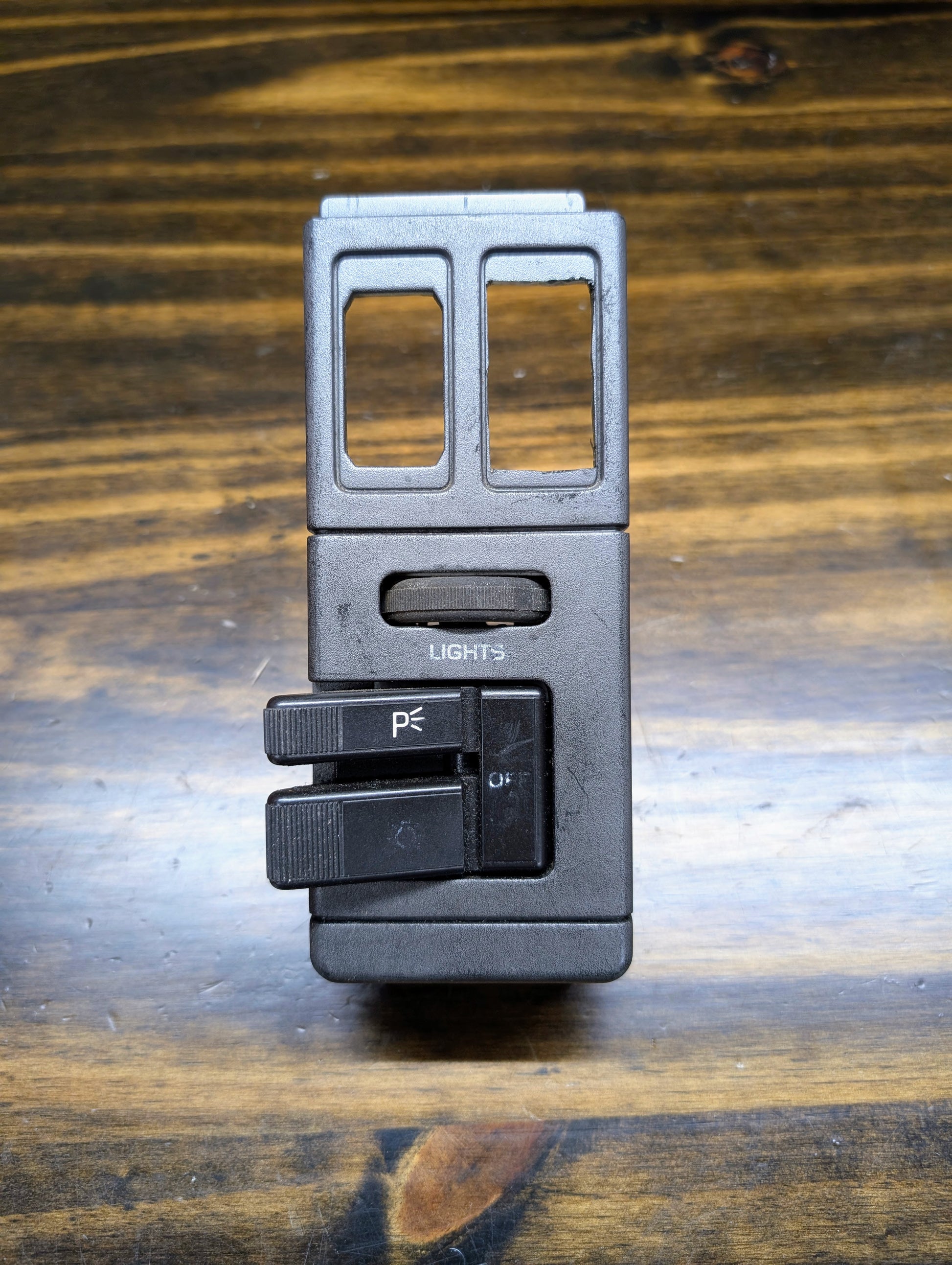 A black OEM General Motors headlight switch for 1986-1994 Chevy S10, Blazer, GMC S15, and Jimmy features two levers marked P and OFF. Ideal replacement part labeled LIGHTS. Shown on a wooden surface.