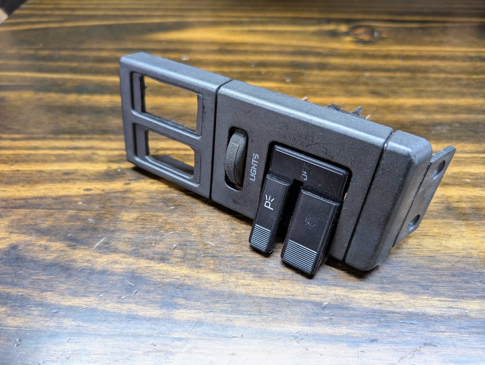 A General Motors OEM Headlight Switch for 1986–1994 Chevy S10, Blazer, GMC S15, and Jimmy sits on wood. The black plastic dashboard assembly features labeled Lights and PR controls with toggle levers and empty rectangular slots.