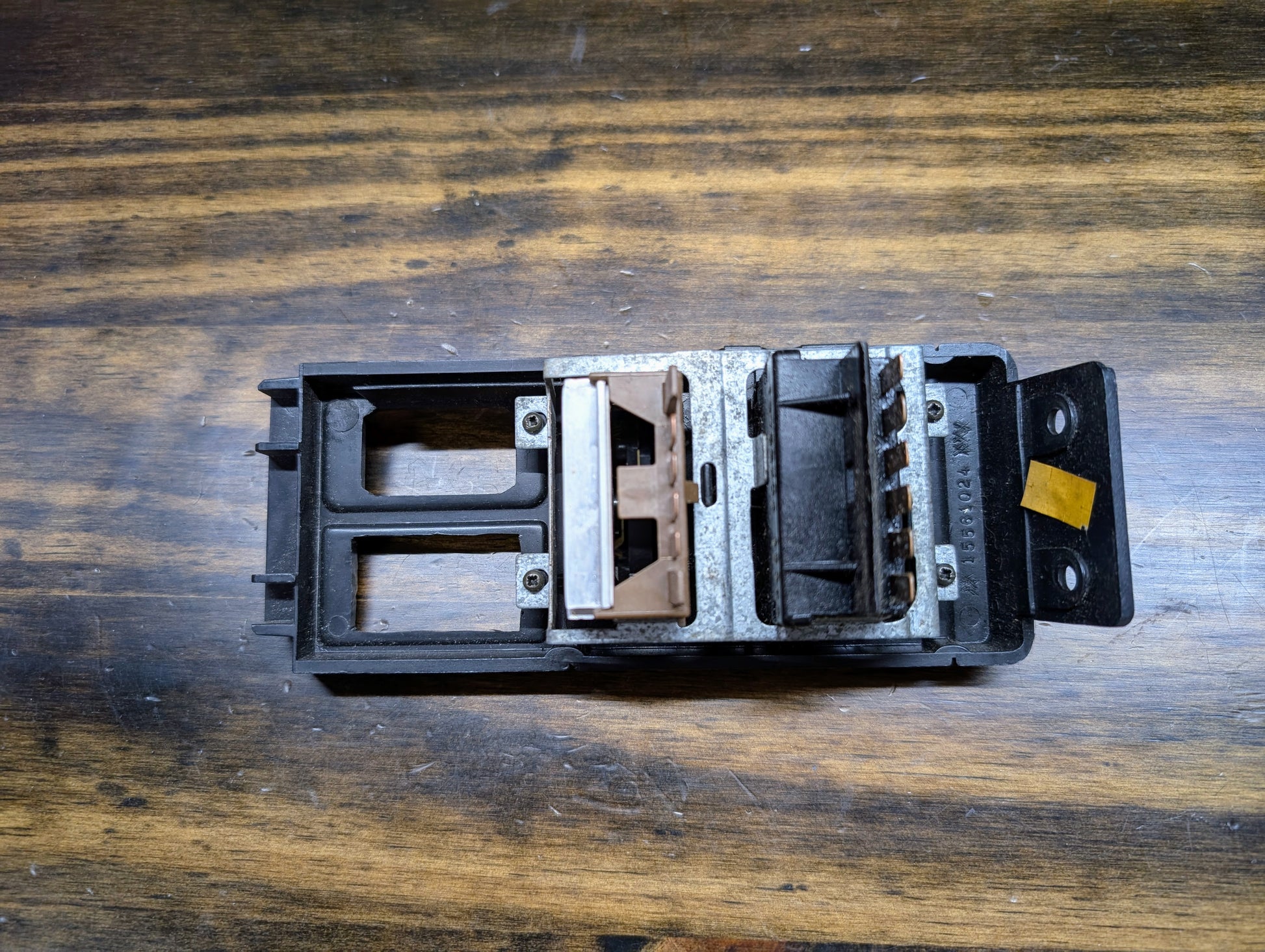 A close-up of a General Motors OEM Headlight Headlamp Switch for 1986-1994 Chevy S10, Blazer, GMC S15, and Jimmy, mounted on a metal and plastic base, shown on a wood surface with visible grain patterns.
