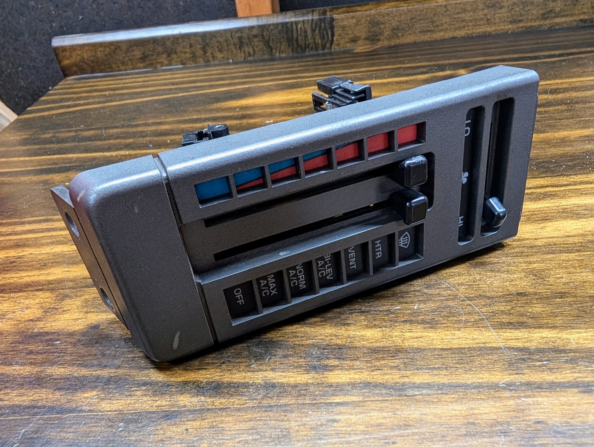 A General Motors HVAC Heater Control Panel with A/C for 1986-1994 Chevrolet S10, Blazer, and more is shown on a wooden surface. The black plastic unit features sliding blue/red levers and labeled airflow buttons.