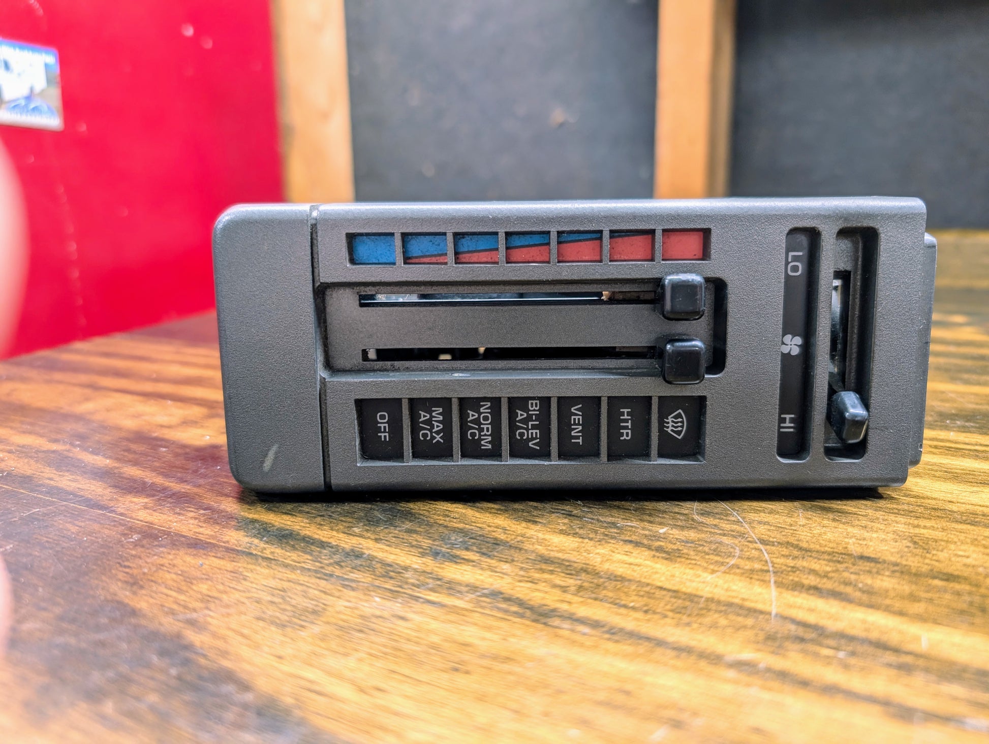 The General Motors HVAC Heater Control Panel with A/C for 1986-1994 Chevrolet S10 and Blazer features sliding levers for temperature and fan speed, labeled airflow buttons, blue/red bars for cool/warm, and a wood-patterned background.