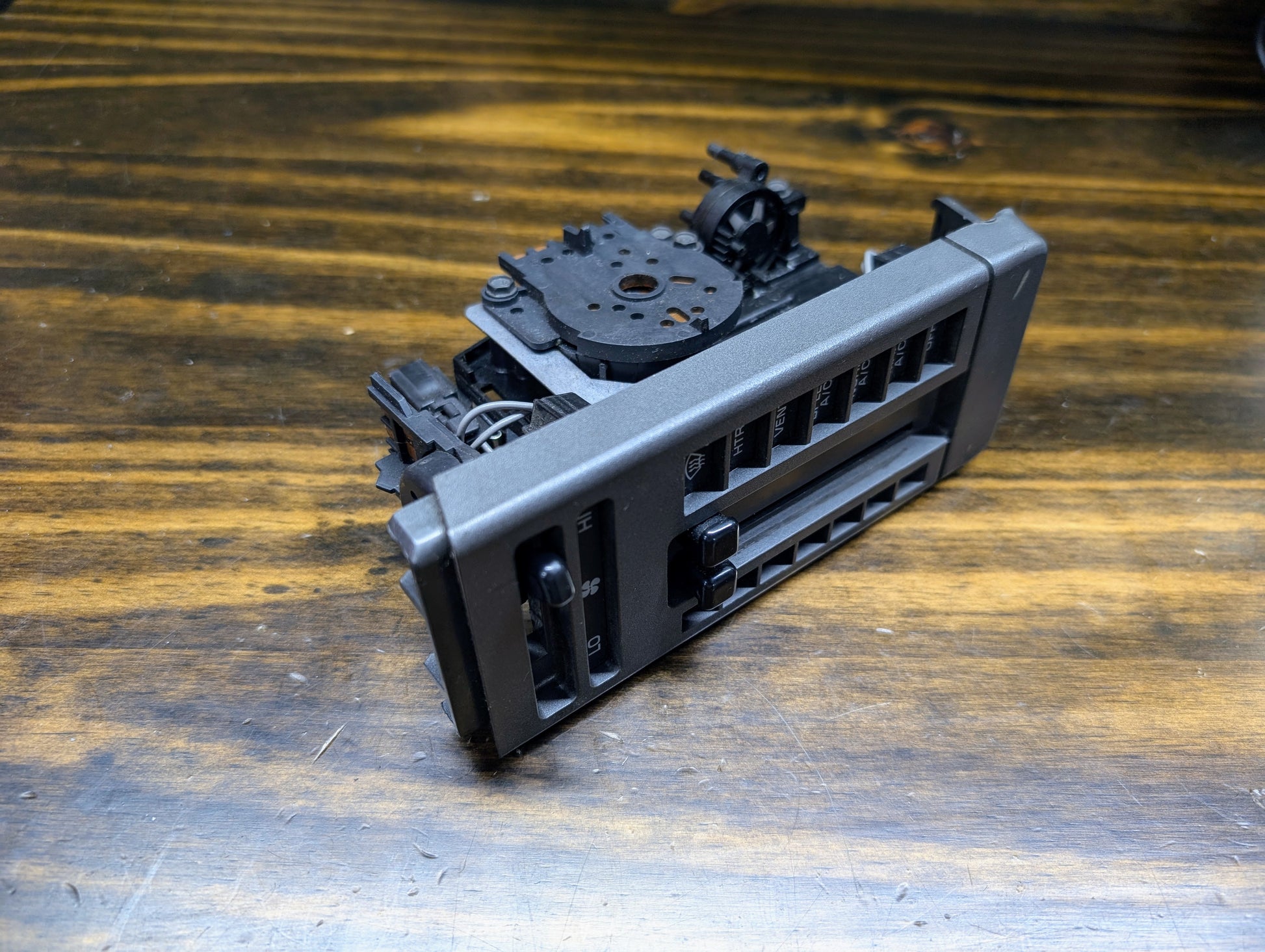 A General Motors HVAC Heater Control Panel with A/C for 1986-1994 Chevrolet S10 and Blazer, along with a dashboard air vent, is shown on a wooden surface with visible internal components and detached parts.