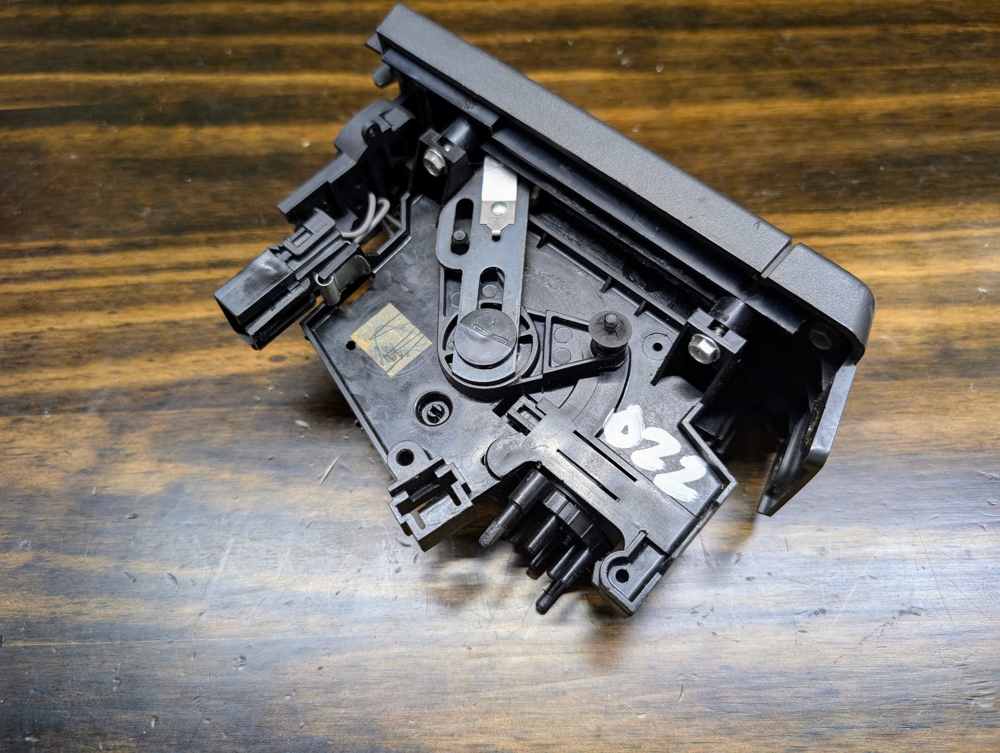 A close-up of a General Motors HVAC Heater Control Panel with A/C for 1986-1994 Chevrolet S10, Blazer, and more rests on a wooden surface. The black plastic part labeled 022 features visible gears and levers.