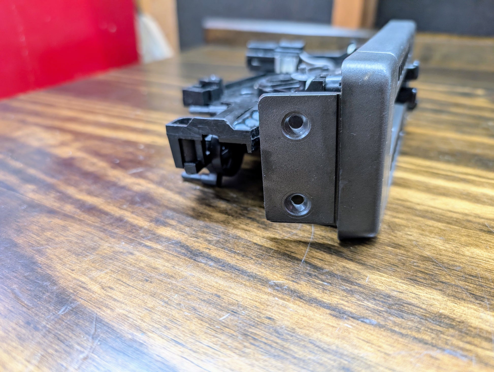 A close-up of a black plastic and metal mounting bracket with two screw holes, part of the General Motors HVAC Heater Control Panel with A/C for 1986-1994 Chevrolet S10, Blazer, and more, shown on wood with a blurred red and black background.