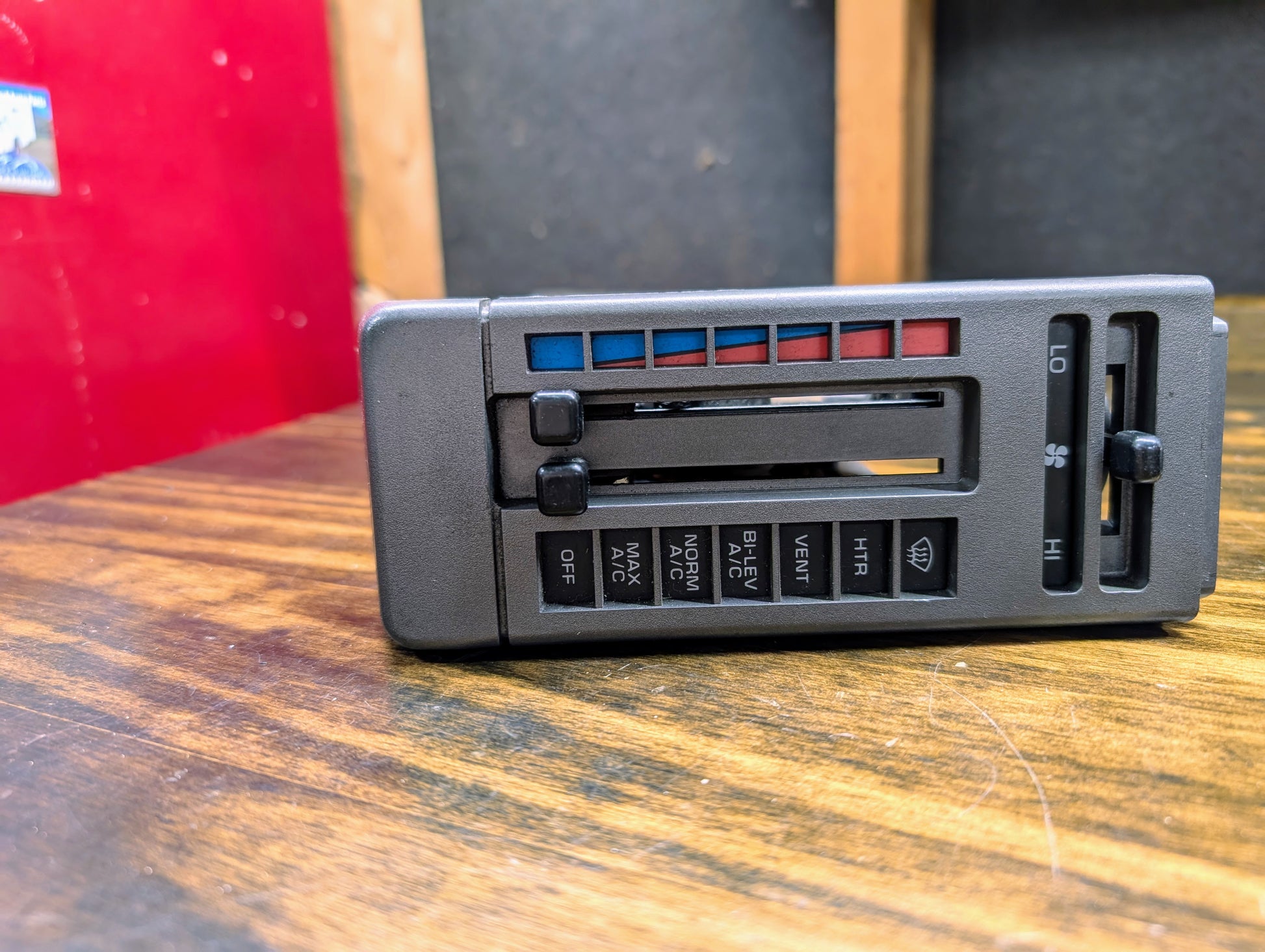 A close-up of a General Motors HVAC Heater Control Panel with A/C for 1986-1994 Chevrolet S10, Blazer, and more, showing sliders for temperature and fan speed, plus buttons for ventilation modes on a wooden surface.