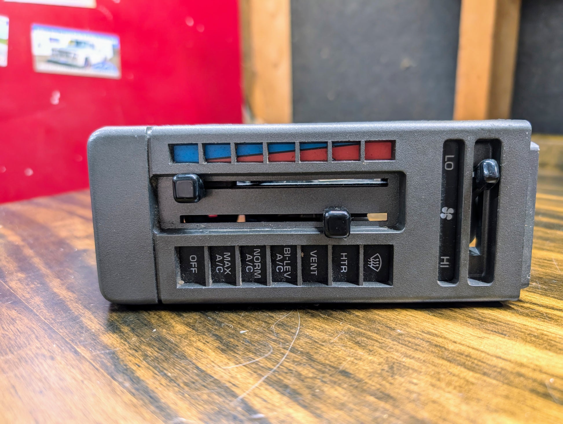 A General Motors HVAC Heater Control Panel with A/C for 1986-1994 Chevrolet S10, Blazer, and more is shown close-up on wood, featuring sliders for temperature and fan speed plus labeled buttons for air conditioning and heating settings.