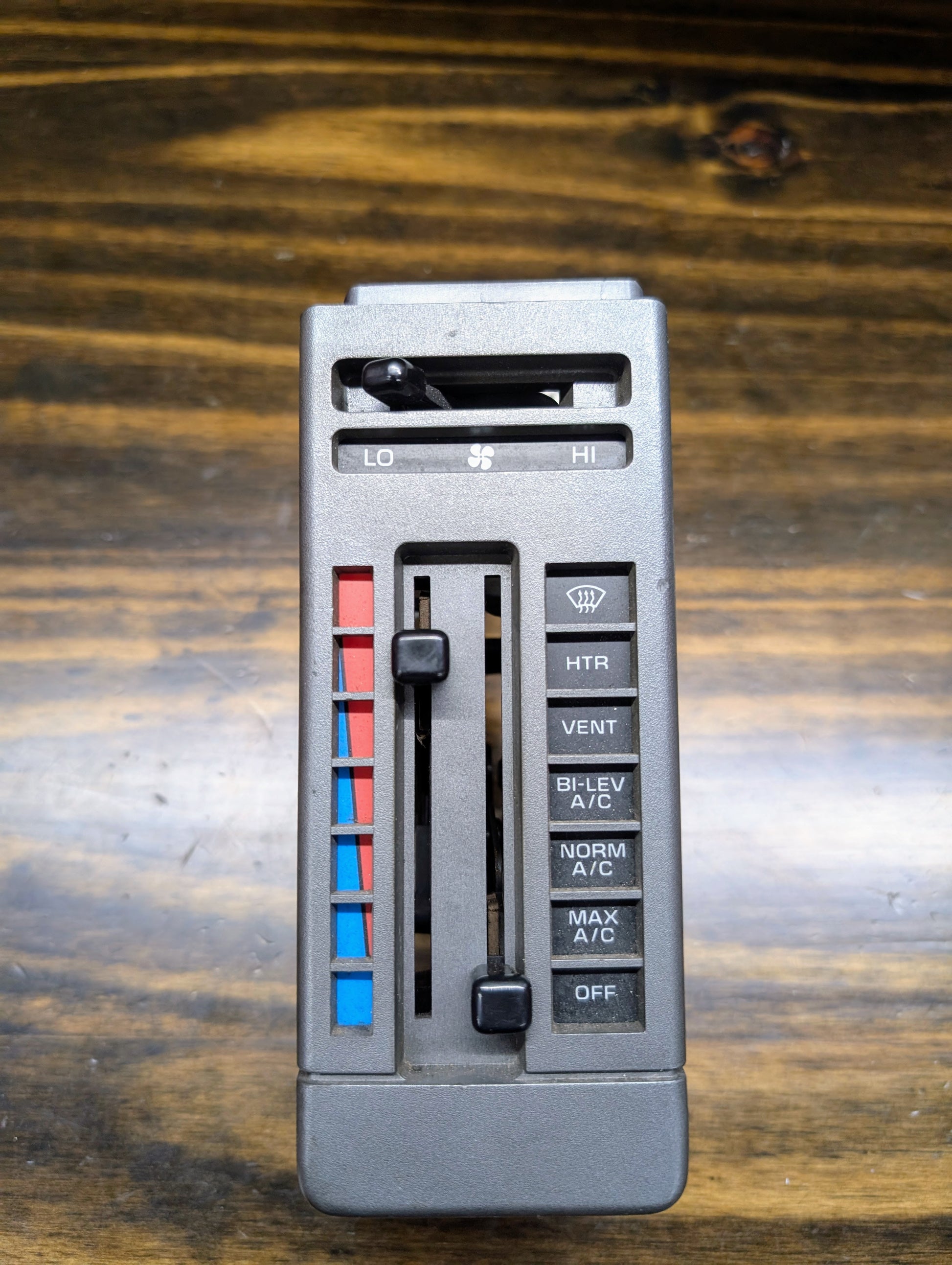 A close-up shows a General Motors HVAC Heater Control Panel with A/C for 1986-1994 Chevrolet S10/Blazer, featuring sliders for temperature and fan speed, blue/red accents, and vent, heater, A/C, defrost, and off buttons on a wooden surface.