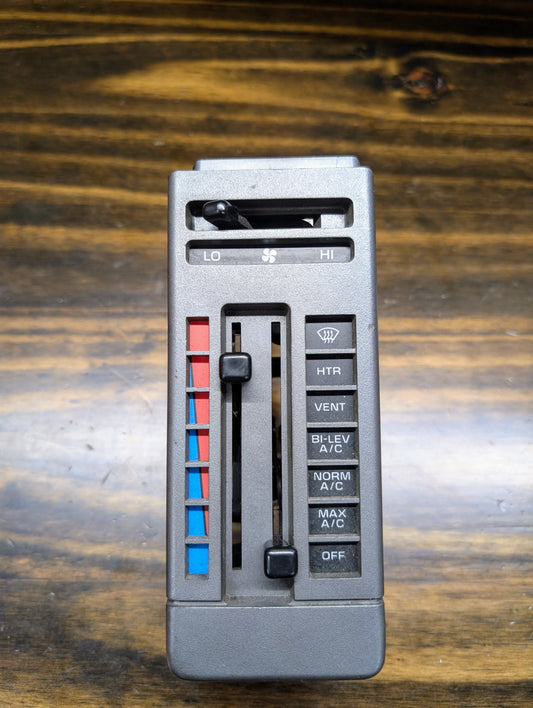 A close-up shows a General Motors HVAC Heater Control Panel with A/C for 1986-1994 Chevrolet S10/Blazer, featuring sliders for temperature and fan speed, blue/red accents, and vent, heater, A/C, defrost, and off buttons on a wooden surface.