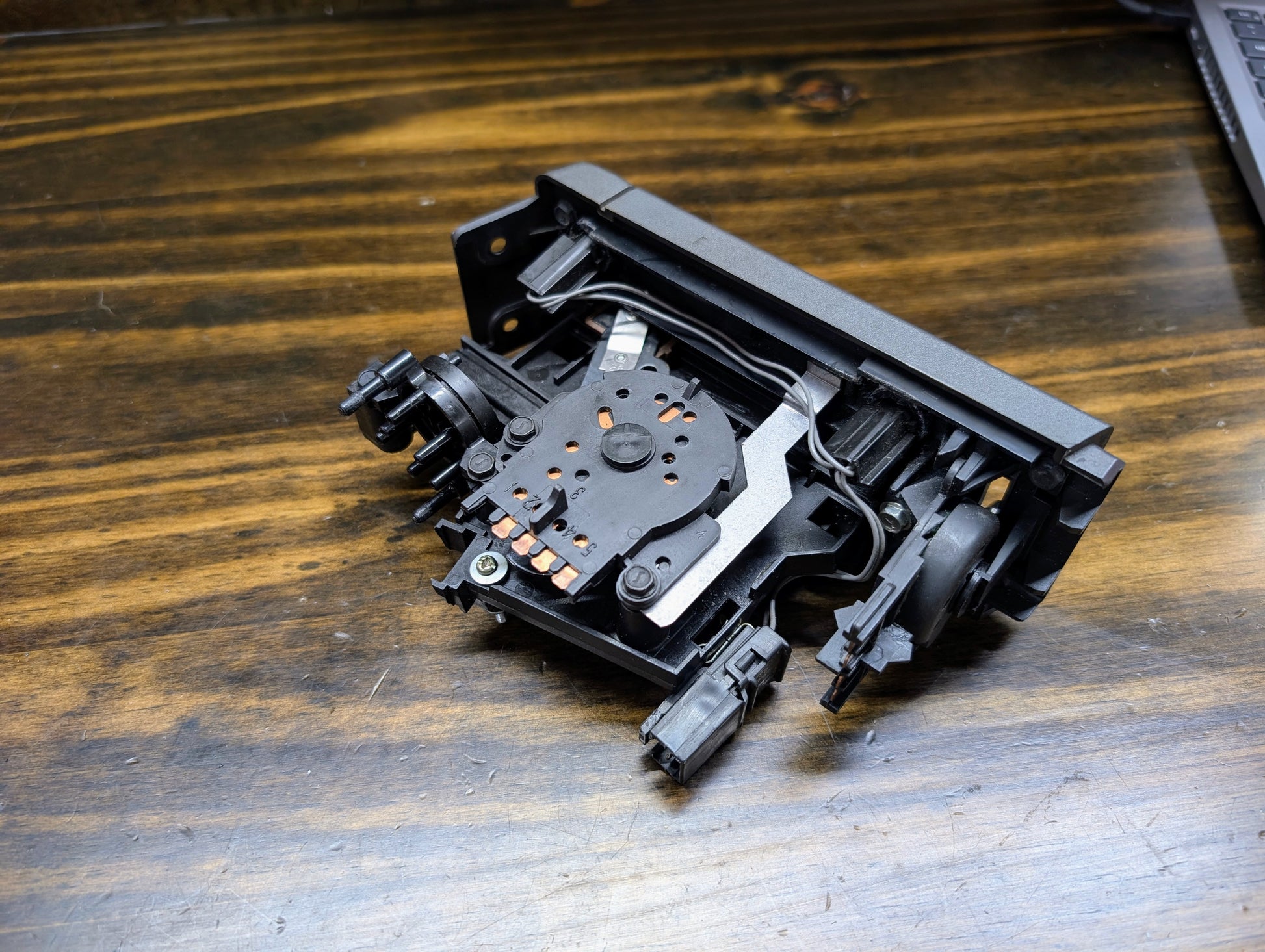 A close-up of a disassembled optical disc drive laser assembly on wood, featuring a General Motors HVAC Heater Control Panel with A/C for 1986-1994 Chevrolet S10, Blazer, and more, plus part of a laptop in the top right.