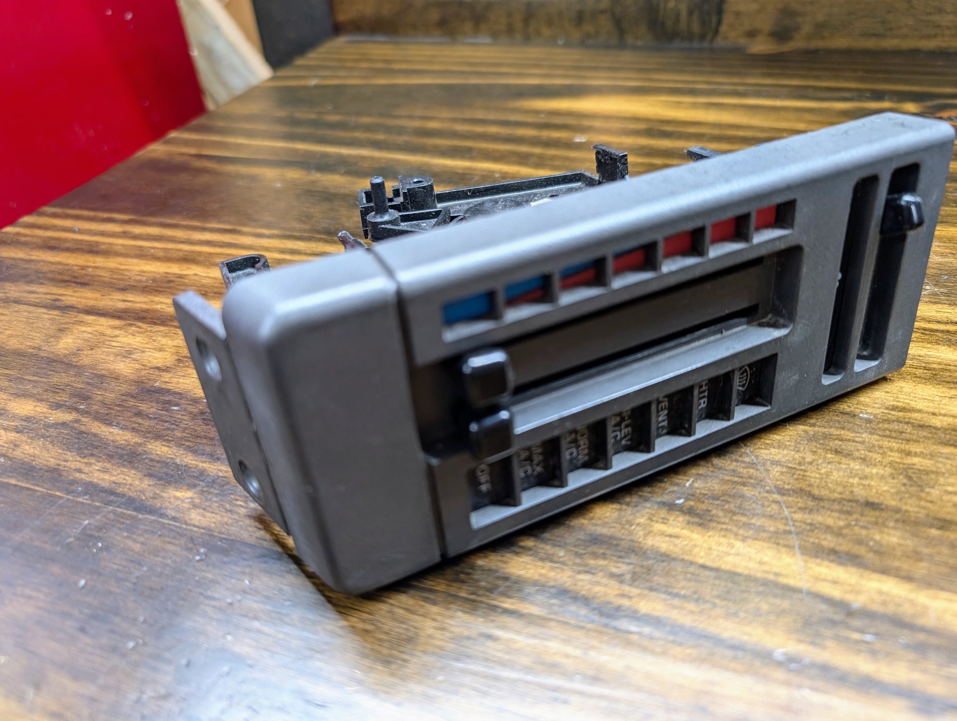 A close-up of a General Motors HVAC Heater Control Panel with A/C for 1986-1994 Chevrolet S10, Blazer, and more, showing sliders, buttons, and blue/red indicators on a wooden surface.