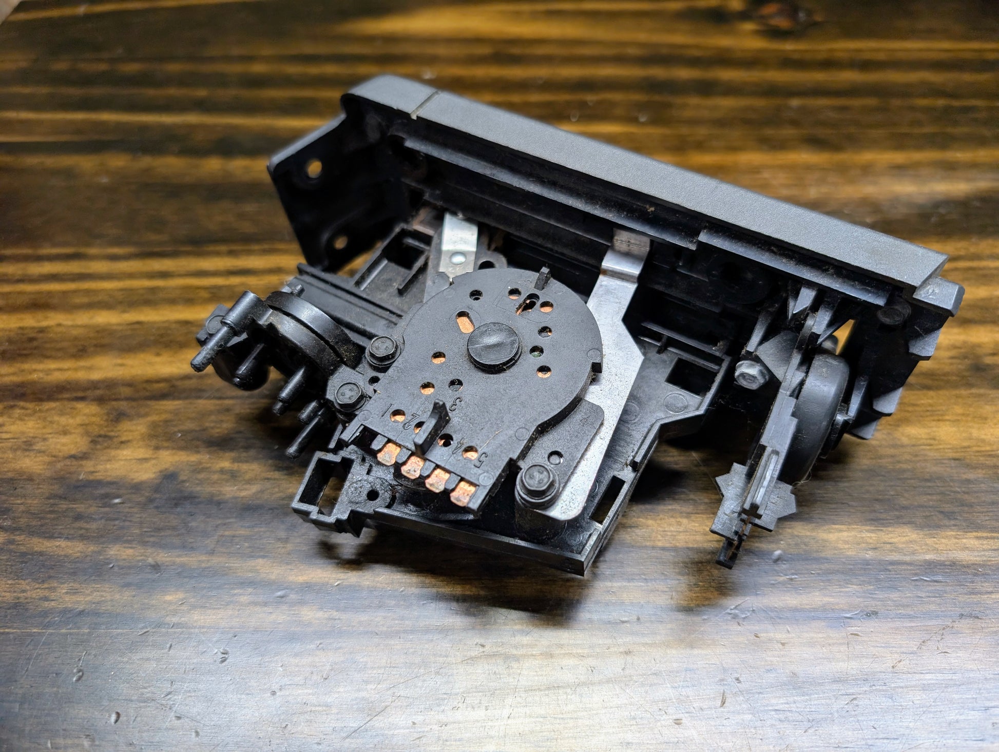 A close-up of a disassembled General Motors HVAC Heater Control Panel with A/C for 1986–1994 Chevrolet S10 and Blazer, displaying copper contacts, gears, and levers on a wooden surface.