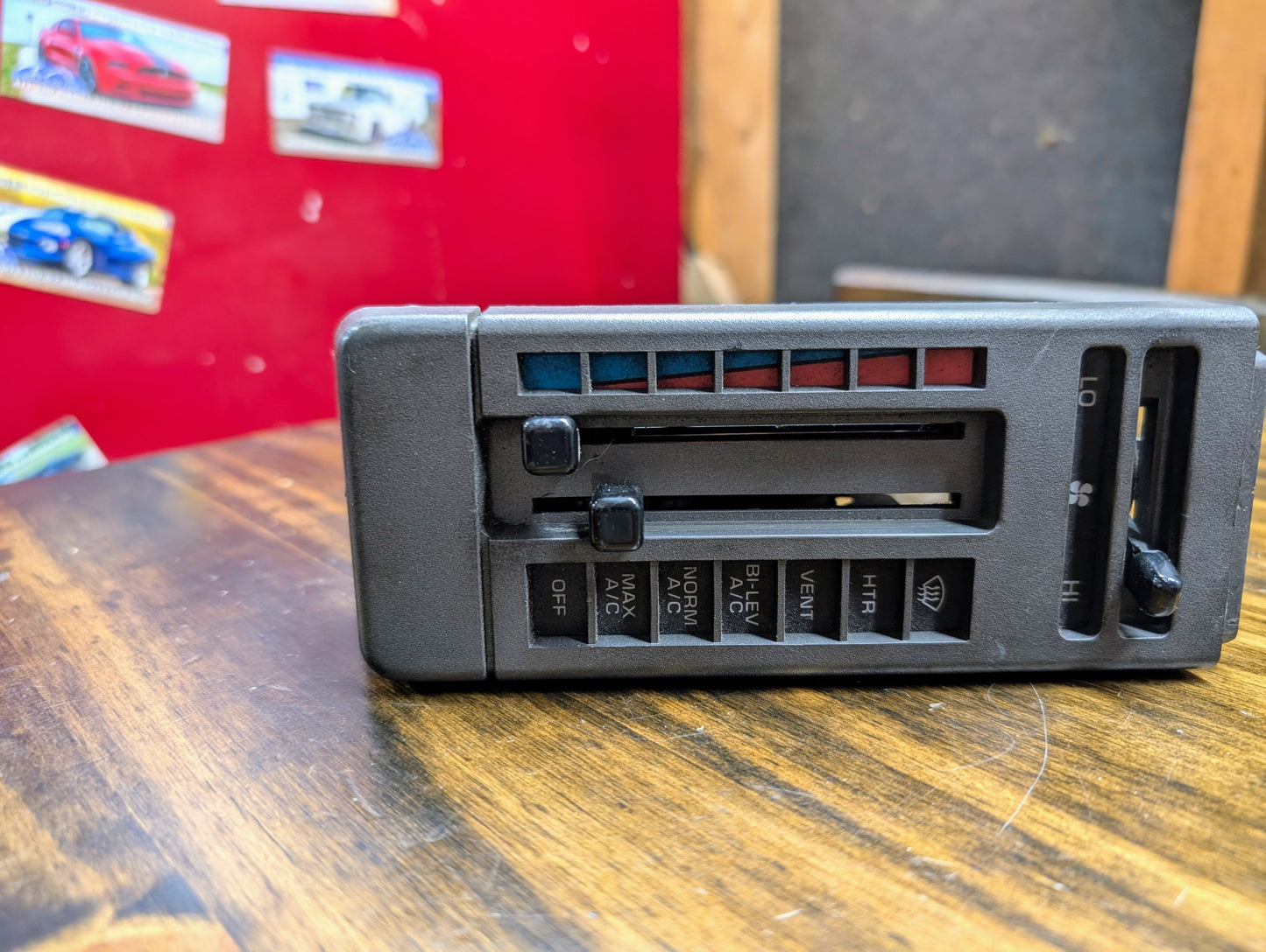 A General Motors HVAC Heater Control Panel with A/C for 1986-1994 Chevrolet S10, Blazer, and more rests on a wooden surface, with red and black walls and car posters in the background.