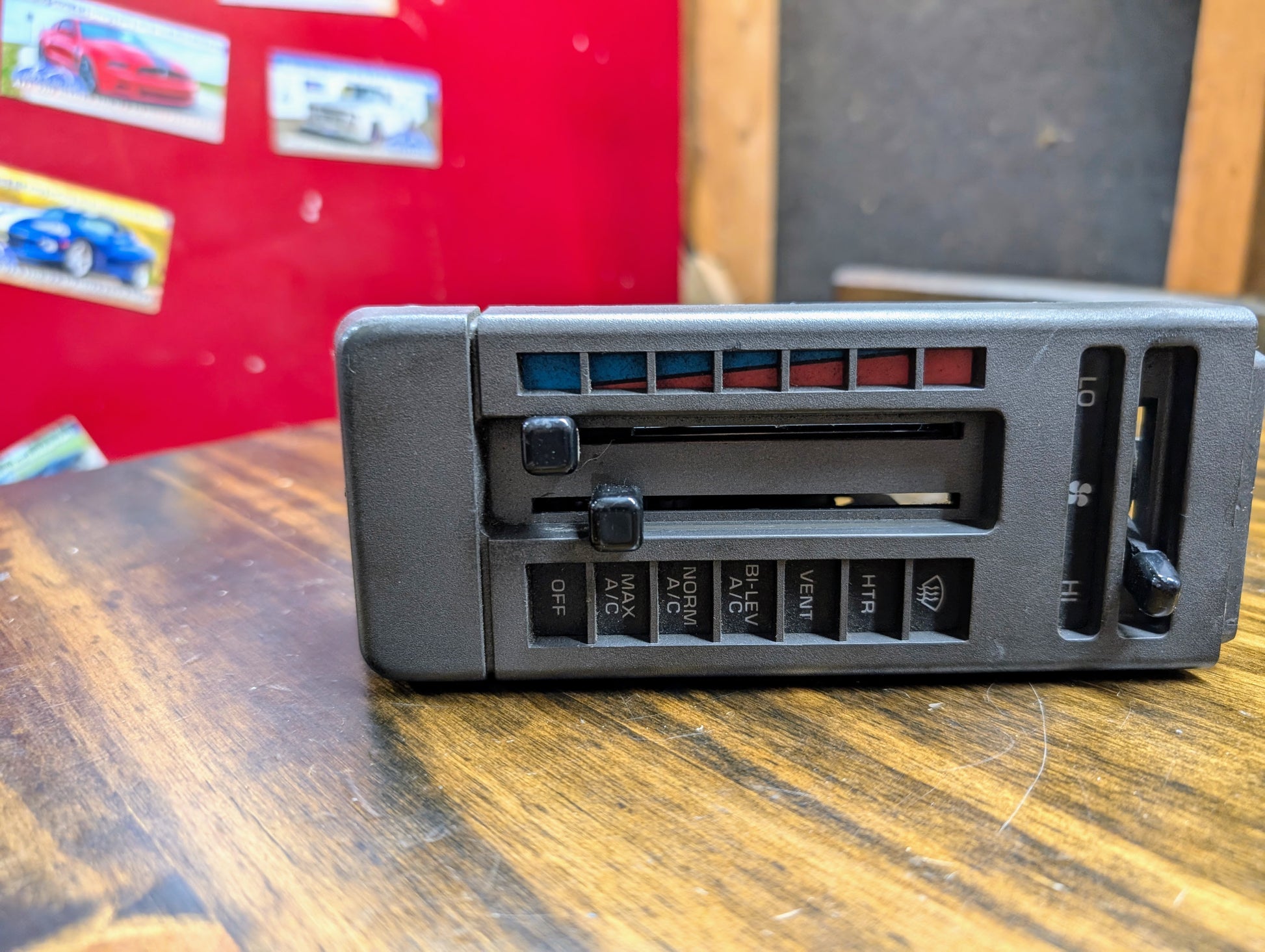 A General Motors HVAC Heater Control Panel with A/C for 1986-1994 Chevrolet S10, Blazer, and more rests on a wooden surface, with red and black walls and car posters in the background.