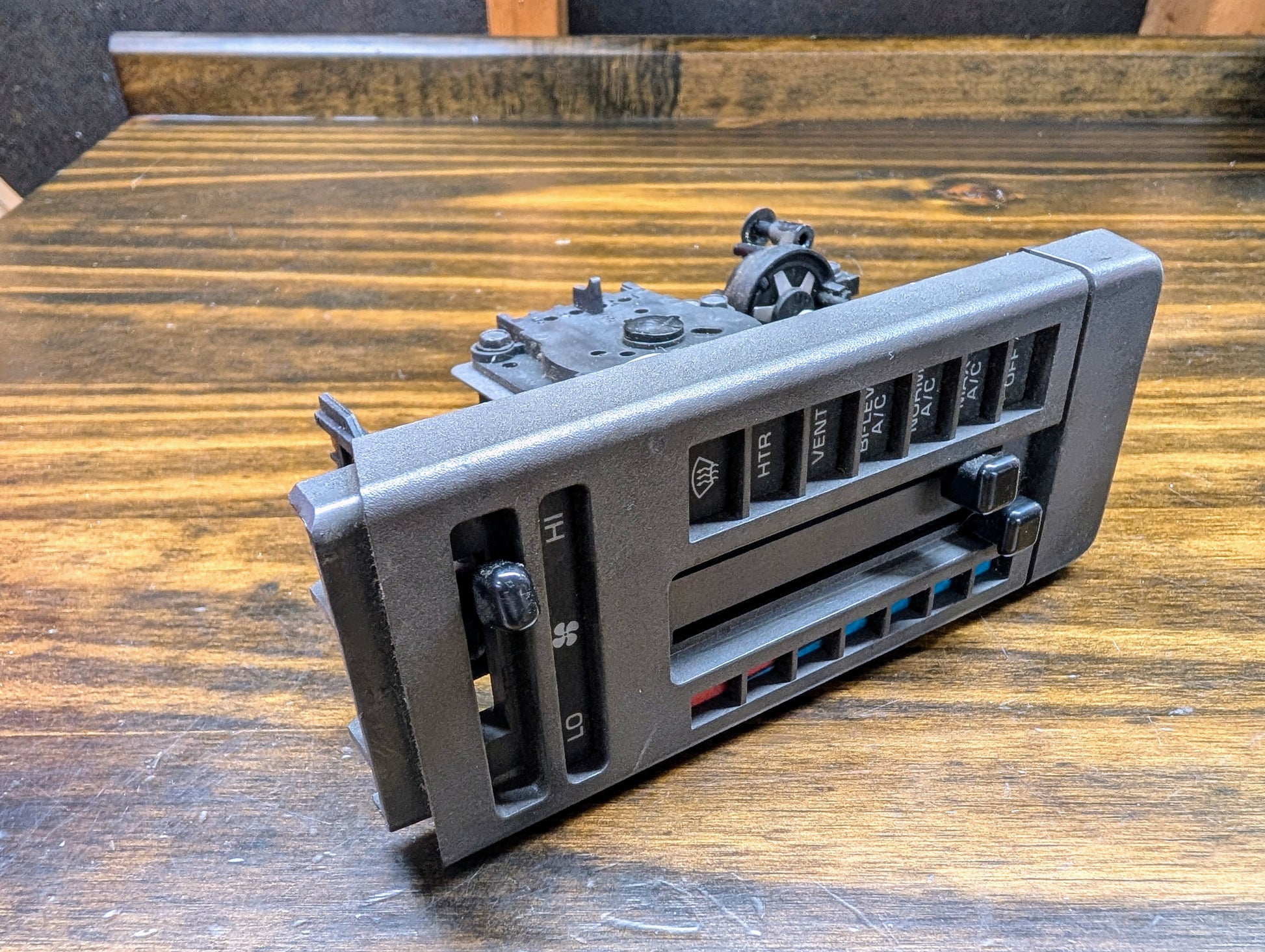 A General Motors HVAC Heater Control Panel with A/C for 1986-1994 Chevrolet S10, Blazer, and more sits on a wooden surface, featuring sliders and buttons to adjust fan speed, temperature, and airflow direction.