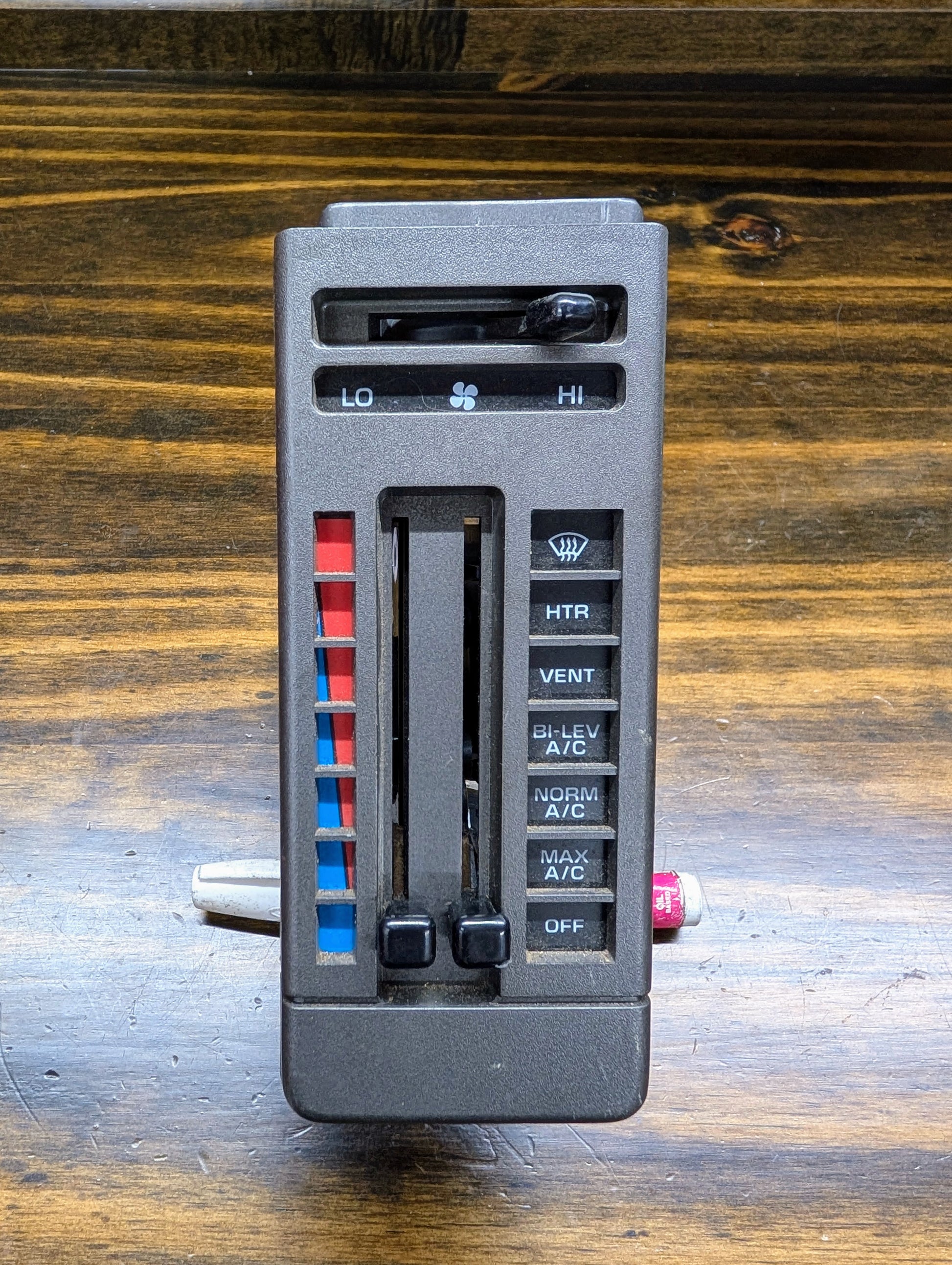 A General Motors HVAC Heater Control Panel with A/C for 1986-1994 Chevrolet S10, Blazer, and more is shown on a wooden surface, featuring sliding levers, blue/red temperature indicators, and labeled heat/vent/defrost/A/C buttons.