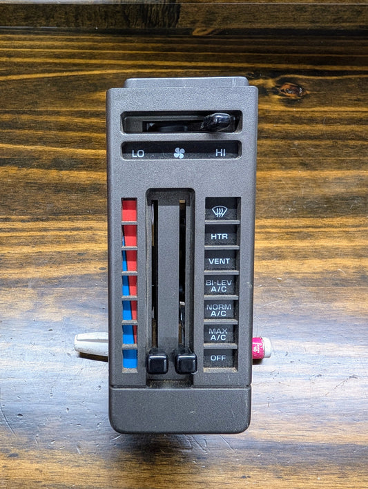 A General Motors HVAC Heater Control Panel with A/C for 1986-1994 Chevrolet S10, Blazer, and more is shown on a wooden surface, featuring sliding levers, blue/red temperature indicators, and labeled heat/vent/defrost/A/C buttons.
