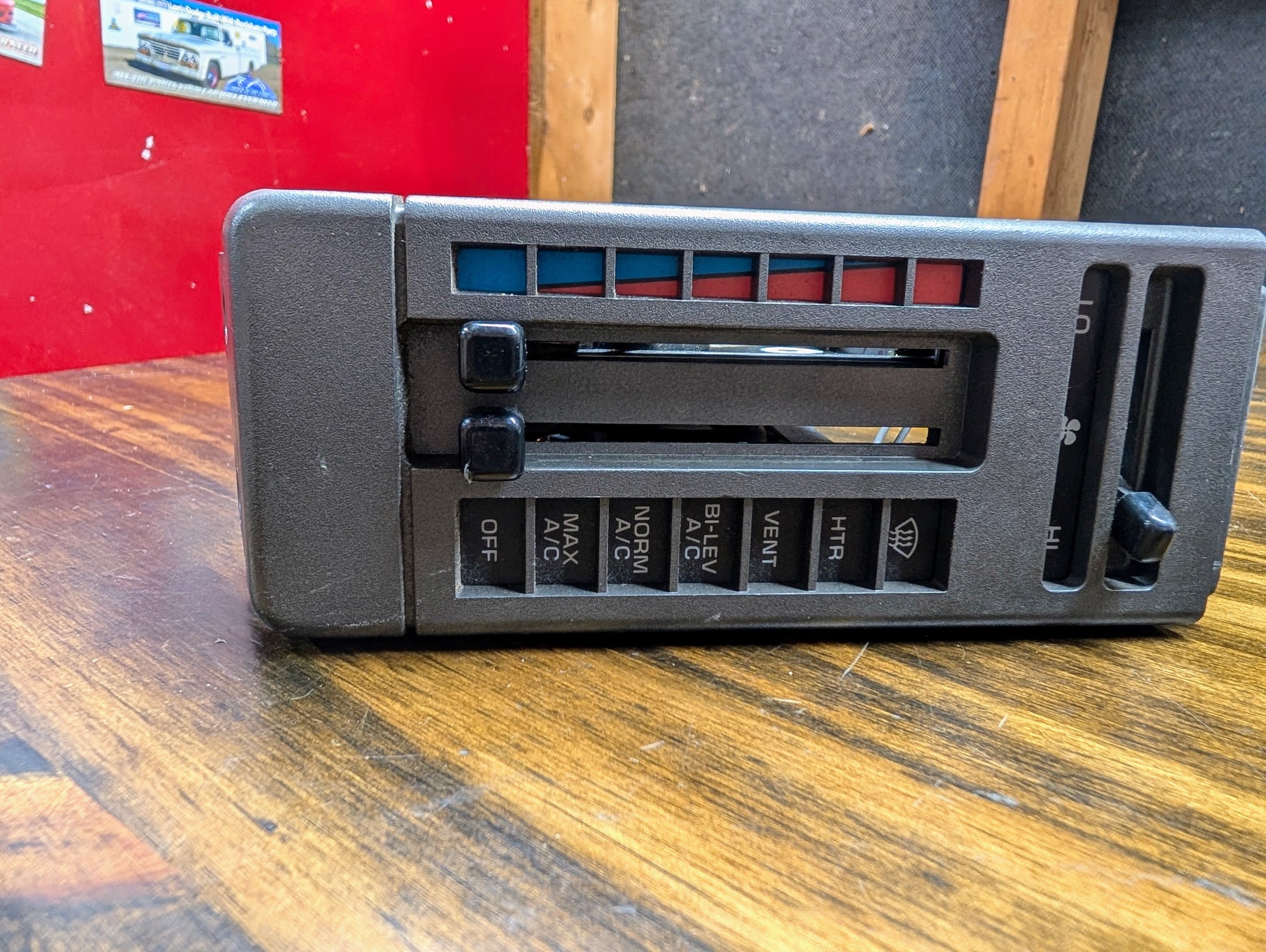 A close-up of the General Motors HVAC Heater Control Panel with A/C for 1986-1994 Chevrolet S10, Blazer, and more, featuring labeled sliders and buttons, displayed on a wooden table in a workshop.