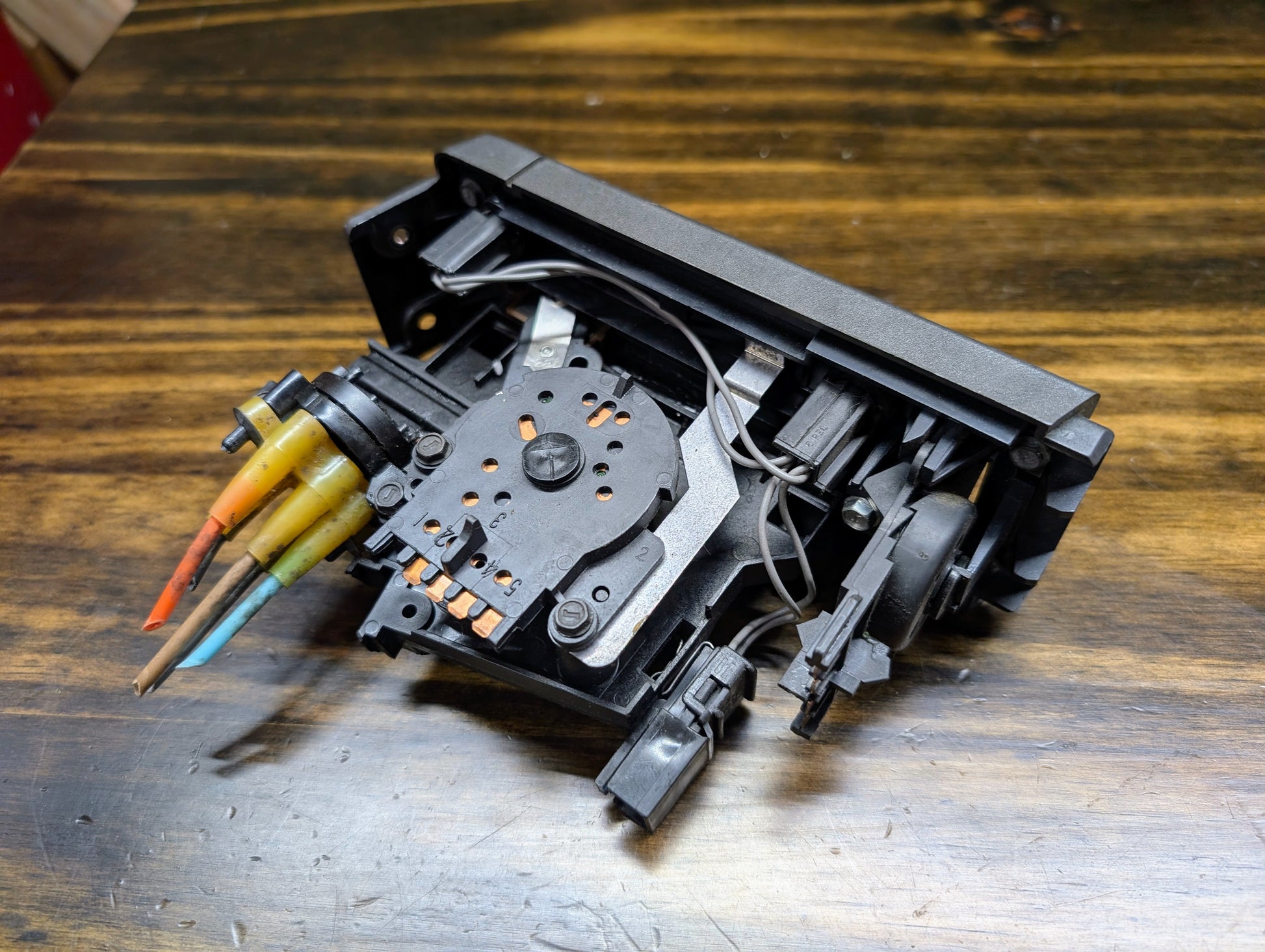 A close-up of a disassembled General Motors HVAC Heater Control Panel with A/C for 1986-1994 Chevrolet S10, Blazer, and more, revealing wires, colored connectors, gears, and metal parts on a wooden surface.
