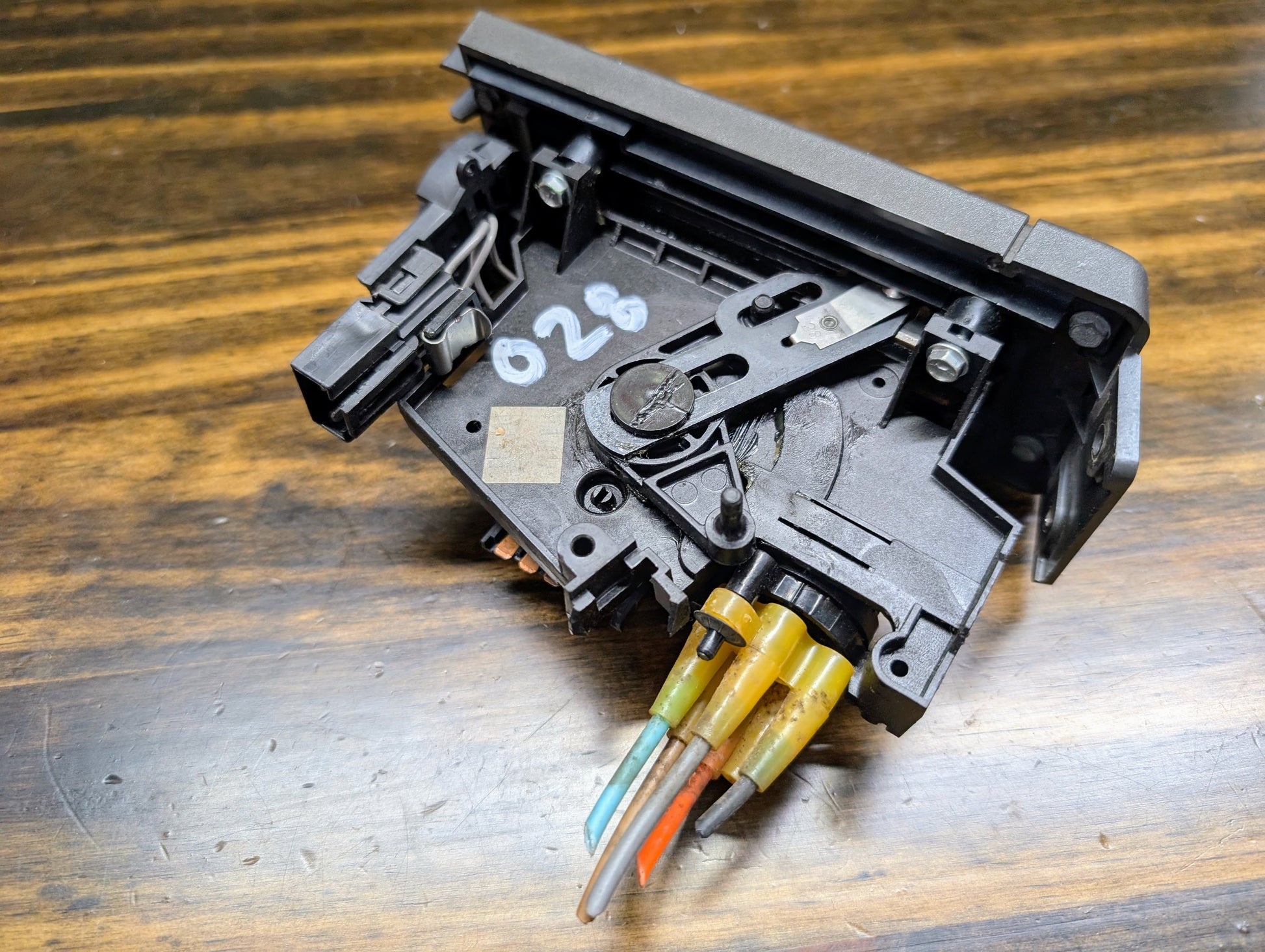 A close-up of a General Motors HVAC Heater Control Panel with A/C for 1986–1994 Chevrolet S10 and Blazer, showing visible wiring—including orange, blue, and green wires—extending from the bottom on a wooden surface.