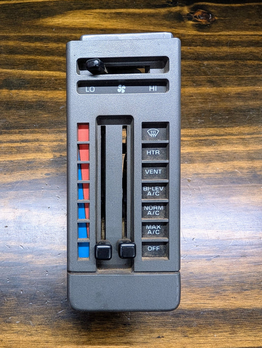 A General Motors HVAC Heater Control Panel with A/C for 1986-1994 Chevrolet S10, Blazer, and more is shown on a wood surface. It features sliders and buttons for temperature, fan speed, heat, vent, bi-level A/C, norm A/C, max A/C, and off.