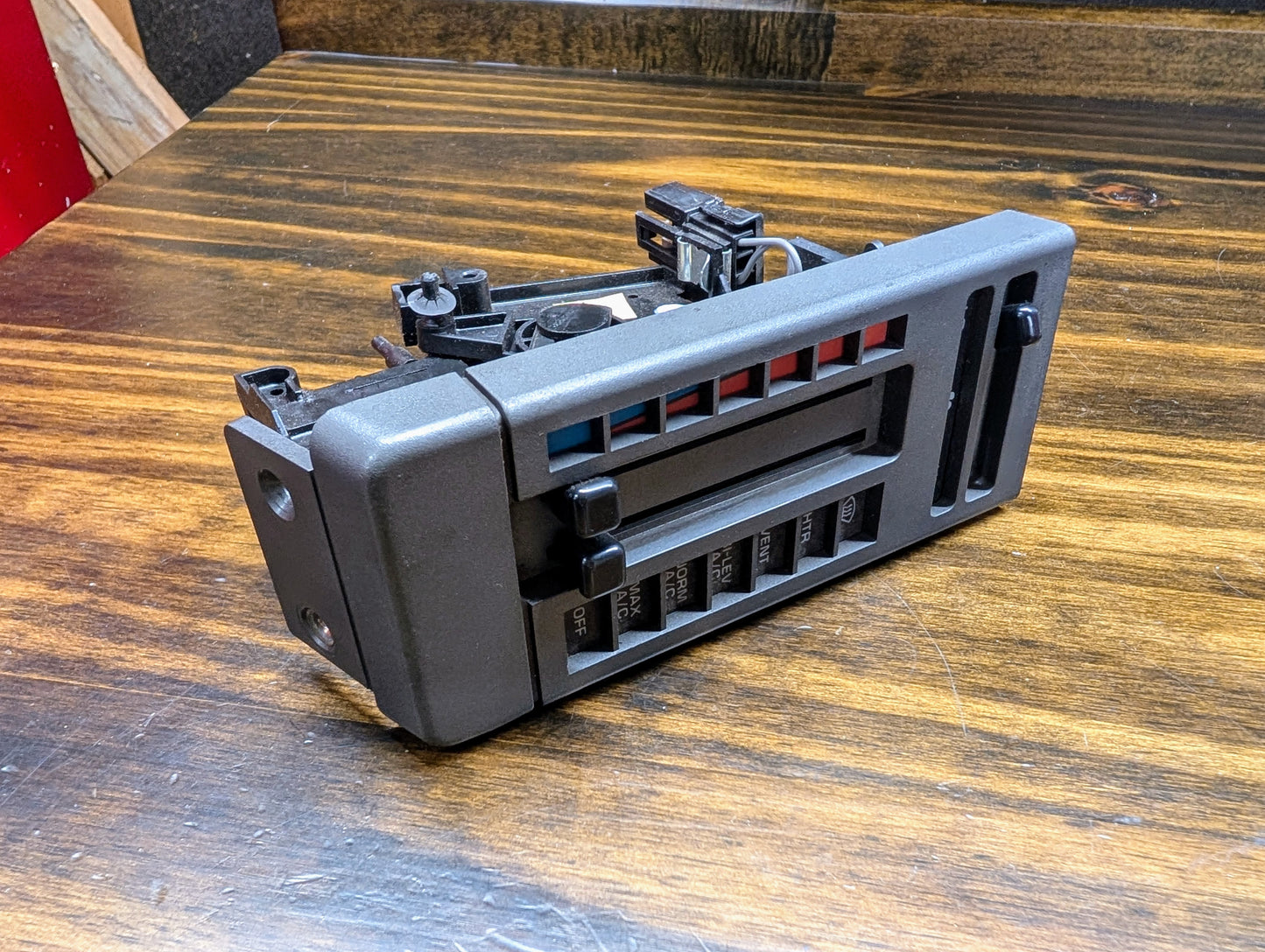 A General Motors HVAC Heater Control Panel with A/C for 1986-1994 Chevrolet S10 or Blazer is shown on a wood surface, featuring black plastic sliders for temperature, airflow, and fan speed with colored hot/cold indicators.