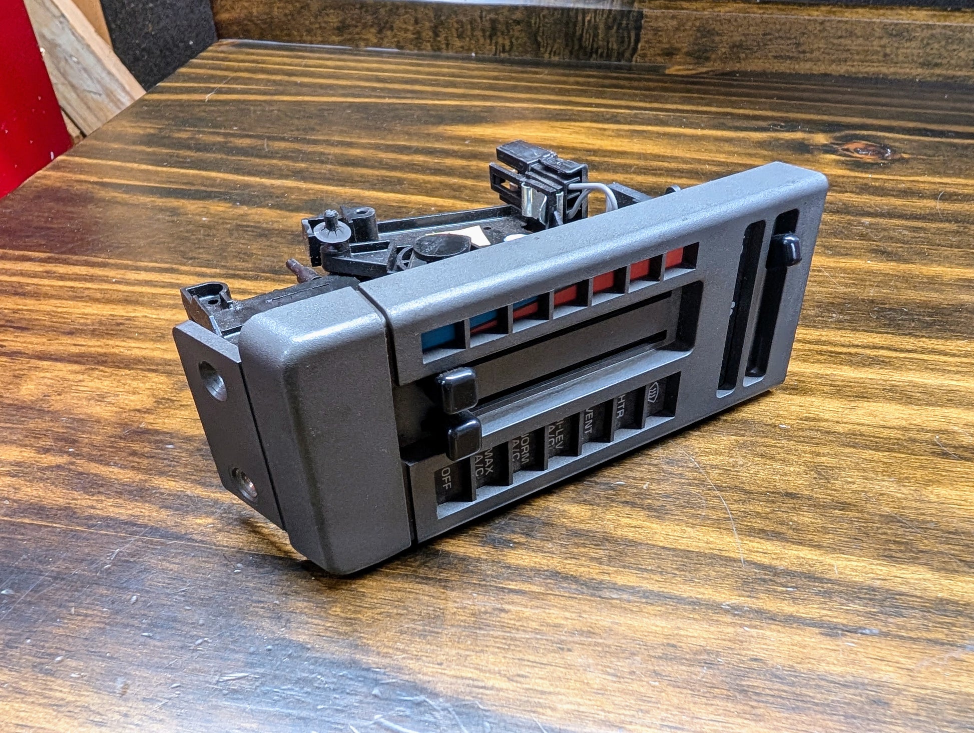 A General Motors HVAC Heater Control Panel with A/C for 1986-1994 Chevrolet S10 or Blazer is shown on a wood surface, featuring black plastic sliders for temperature, airflow, and fan speed with colored hot/cold indicators.