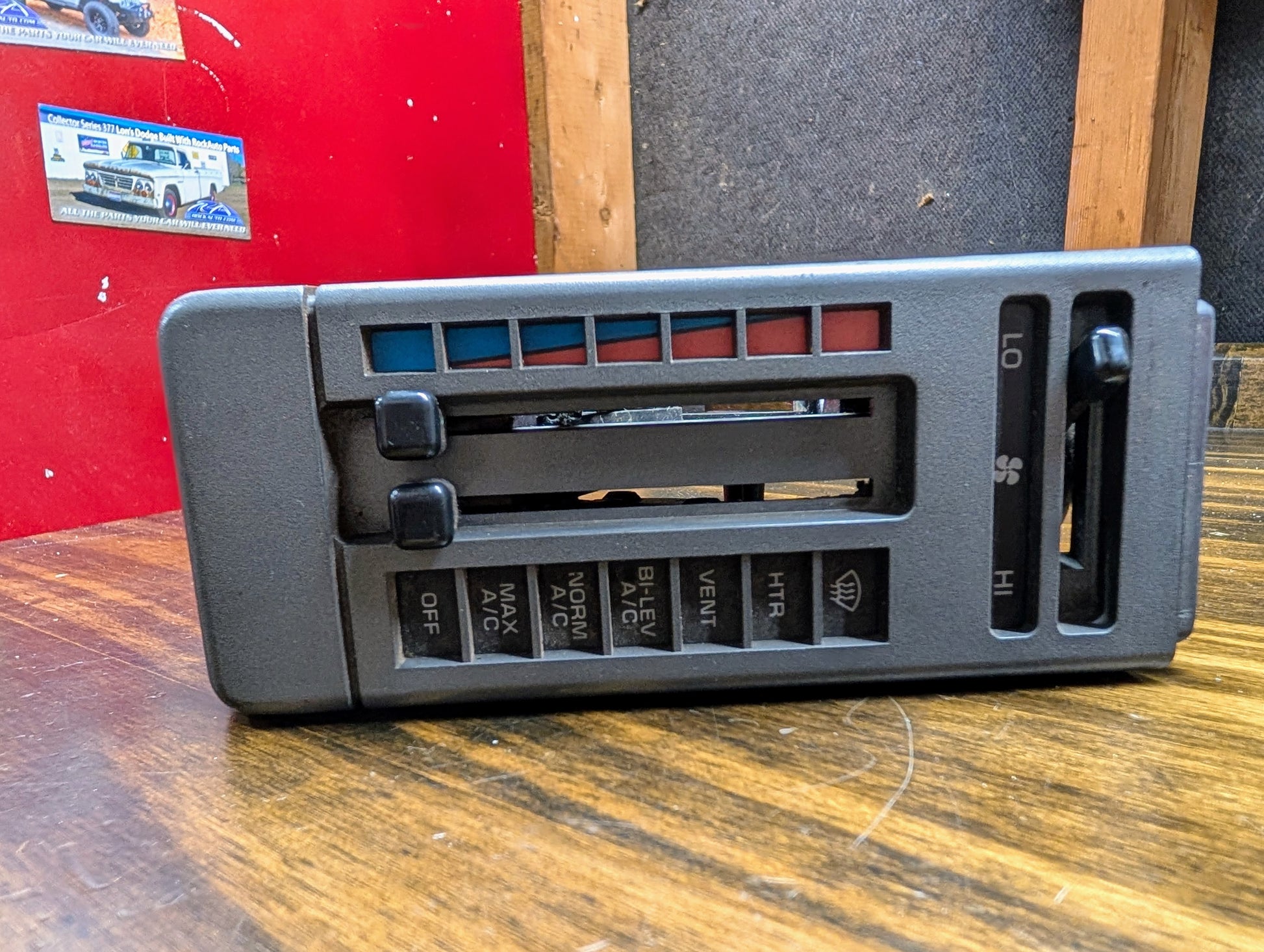 A close-up of a General Motors HVAC Heater Control Panel with A/C for 1986-1994 Chevrolet S10, Blazer, and more shows sliders and buttons for fan speed, vents, temperature, and AC on a wooden surface with a red and gray background.