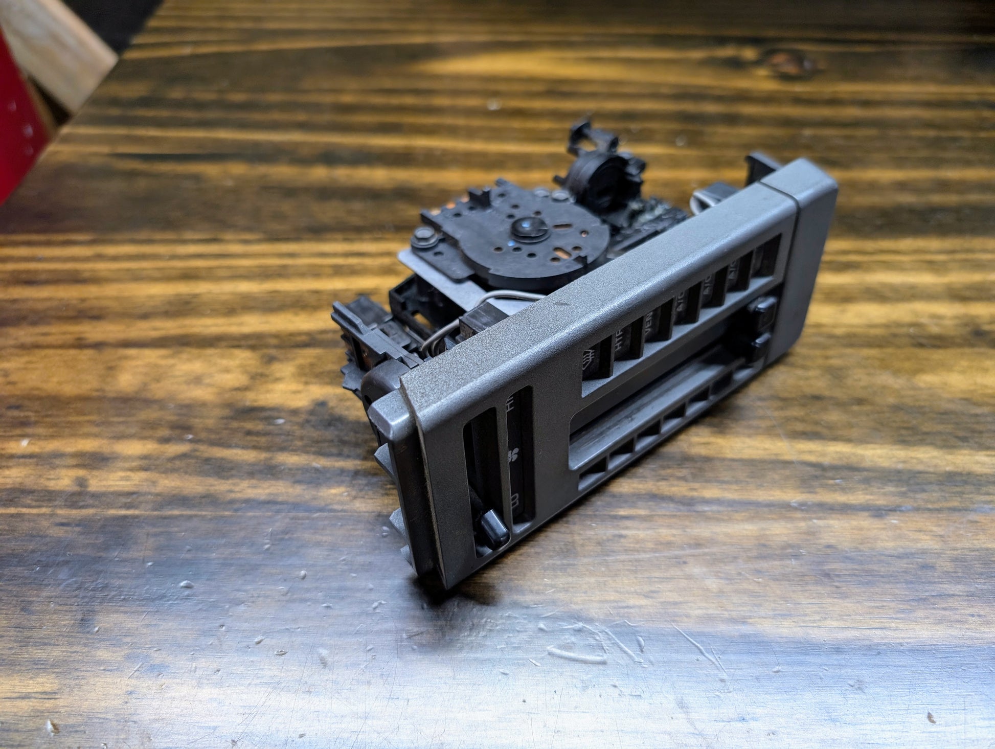 A broken cassette deck with exposed gears sits on a wood-grain surface beside a detached General Motors HVAC Heater Control Panel with A/C for 1986–1994 Chevrolet S10, Blazer, and more.