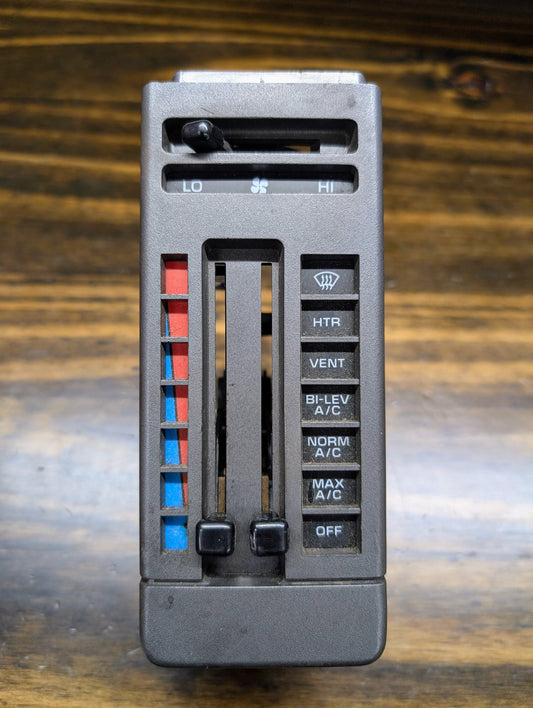 Close-up of a General Motors HVAC Heater Control Panel with A/C for 1986-1994 Chevrolet S10 and Blazer, showing sliders for temperature and fan speed plus buttons for air conditioning and heating on a wooden surface.
