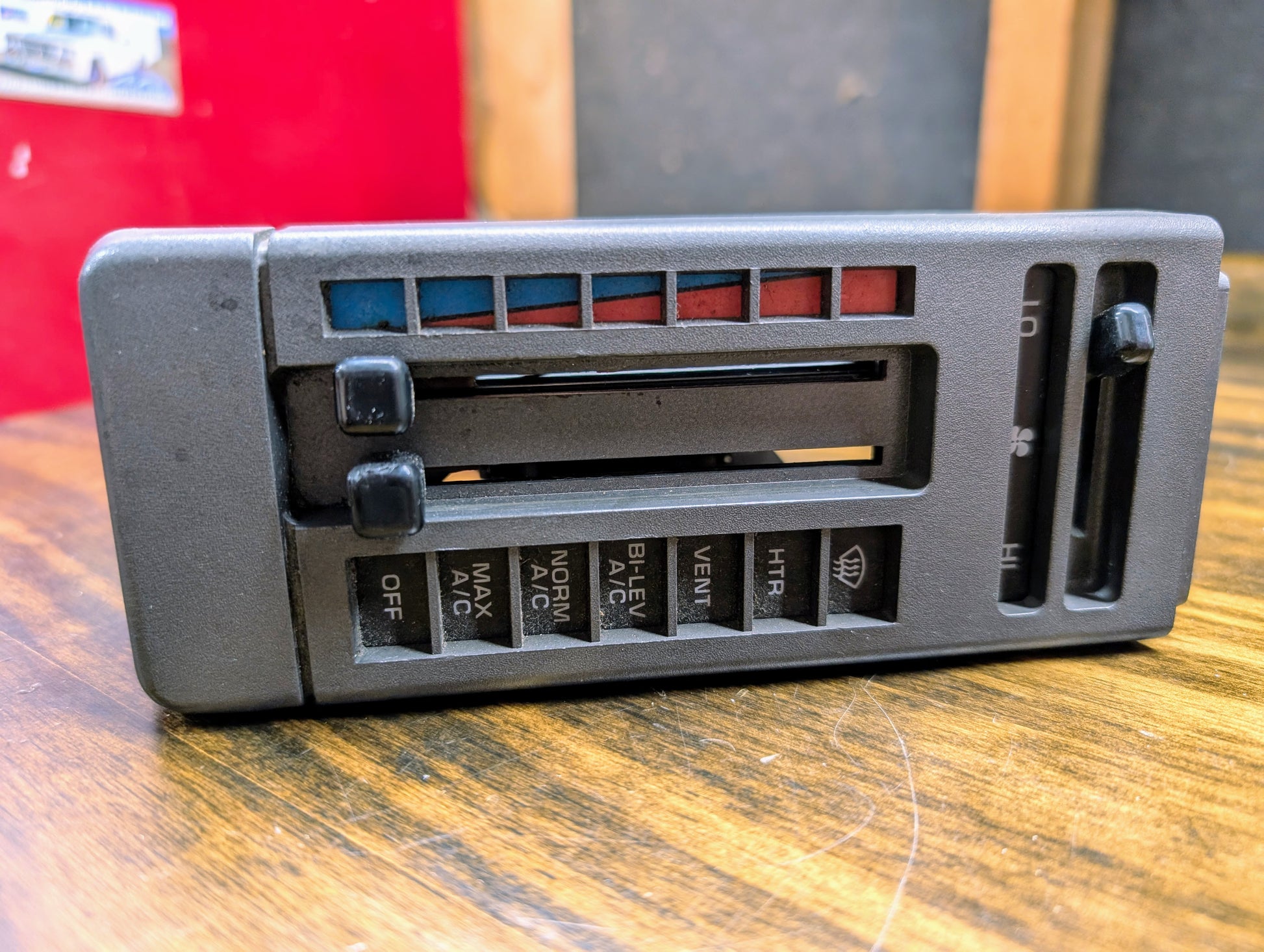A close-up shows the General Motors HVAC Heater Control Panel with A/C for 1986-1994 Chevrolet S10 and Blazer, featuring sliders and labeled buttons for fan speed, temperature, and air flow on a wooden surface.