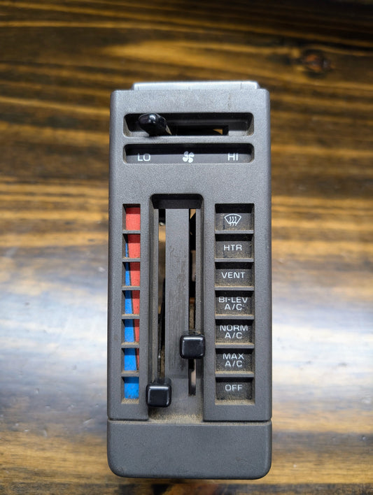 A close-up of the General Motors HVAC Heater Control Panel with A/C for 1986-1994 Chevrolet S10 and Blazer, featuring sliders, blue/red temperature bars, fan speed dial, and heat, vent, A/C, defrost buttons on a wooden background.