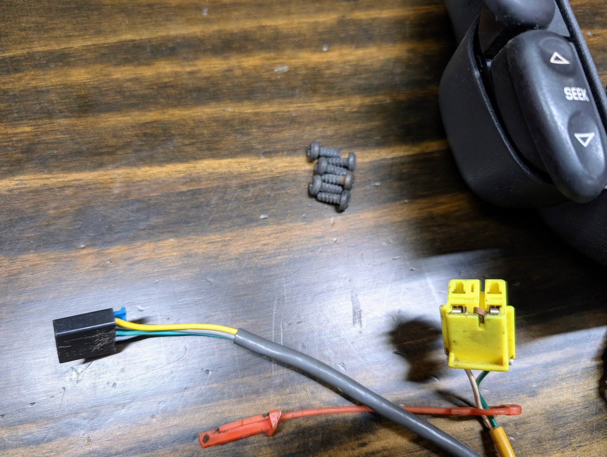 Close-up of General Motors KIT Steering Wheel Black Button Radio Controls & Clock Spring for 2004-2005 Chevy S10, Blazer, and more, plus screws and wires with black/yellow connectors on a wooden surface.