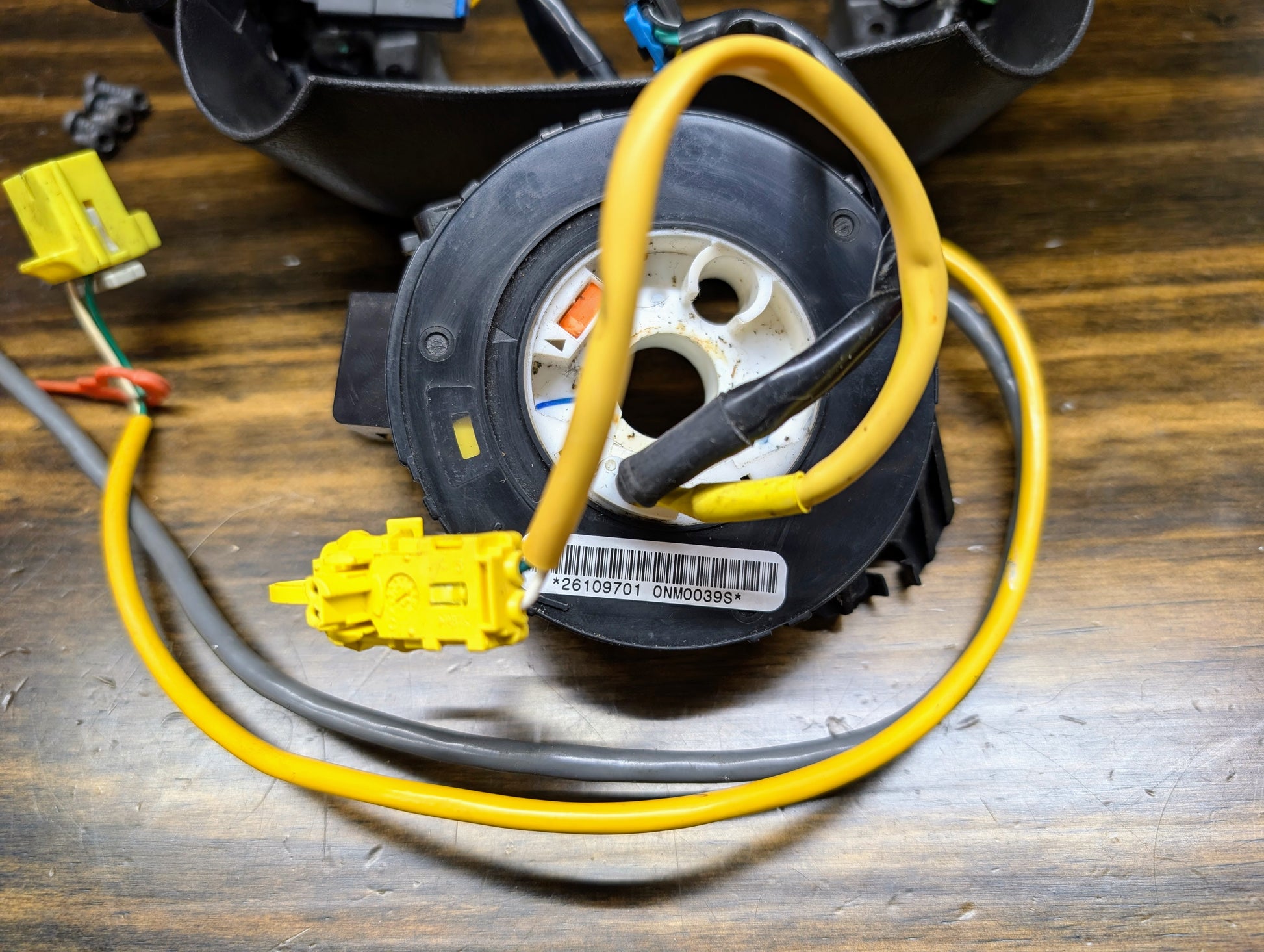 A close-up of the General Motors KIT Steering Wheel Black Button Radio Controls & Clock Spring for 2004-2005 Chevy S10, Blazer, and more, showing attached yellow and gray wires, barcode, connectors, on a wooden surface.