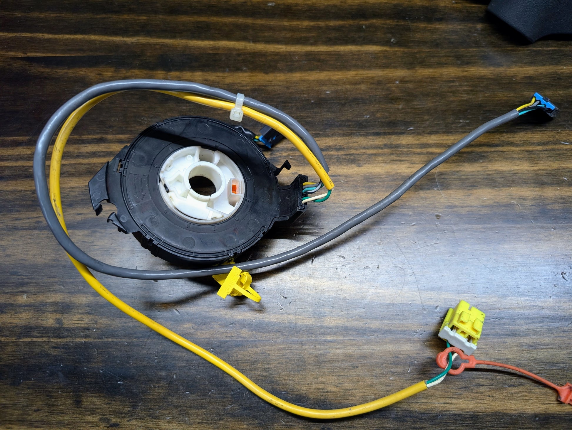 A General Motors KIT Steering Wheel Black Button Radio Controls & Clock Spring for 2004-2005 Chevy S10 and Blazer, with connectors, yellow and grey wires, and zip ties, is displayed on a wooden surface; the round black-and-white unit is central.