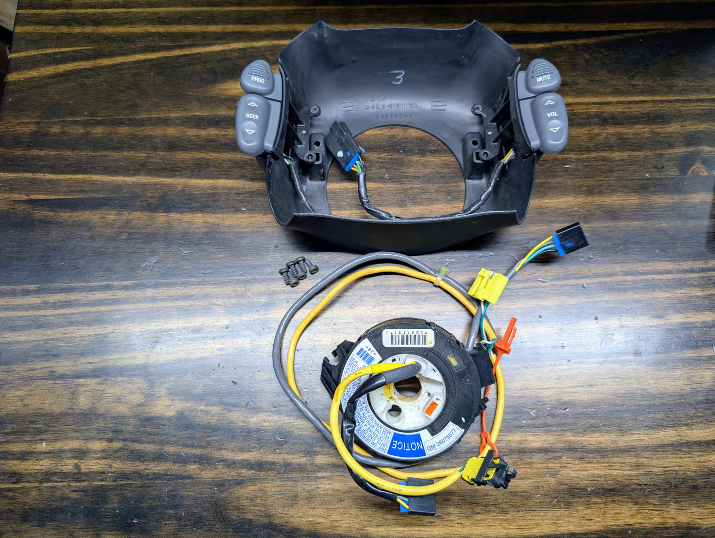 A disassembled General Motors KIT Steering Wheel Radio Controls & Clock Spring for 1999 Chevy S10, Blazer, and more is displayed on a wooden surface, showing cruise and audio buttons plus connectors.