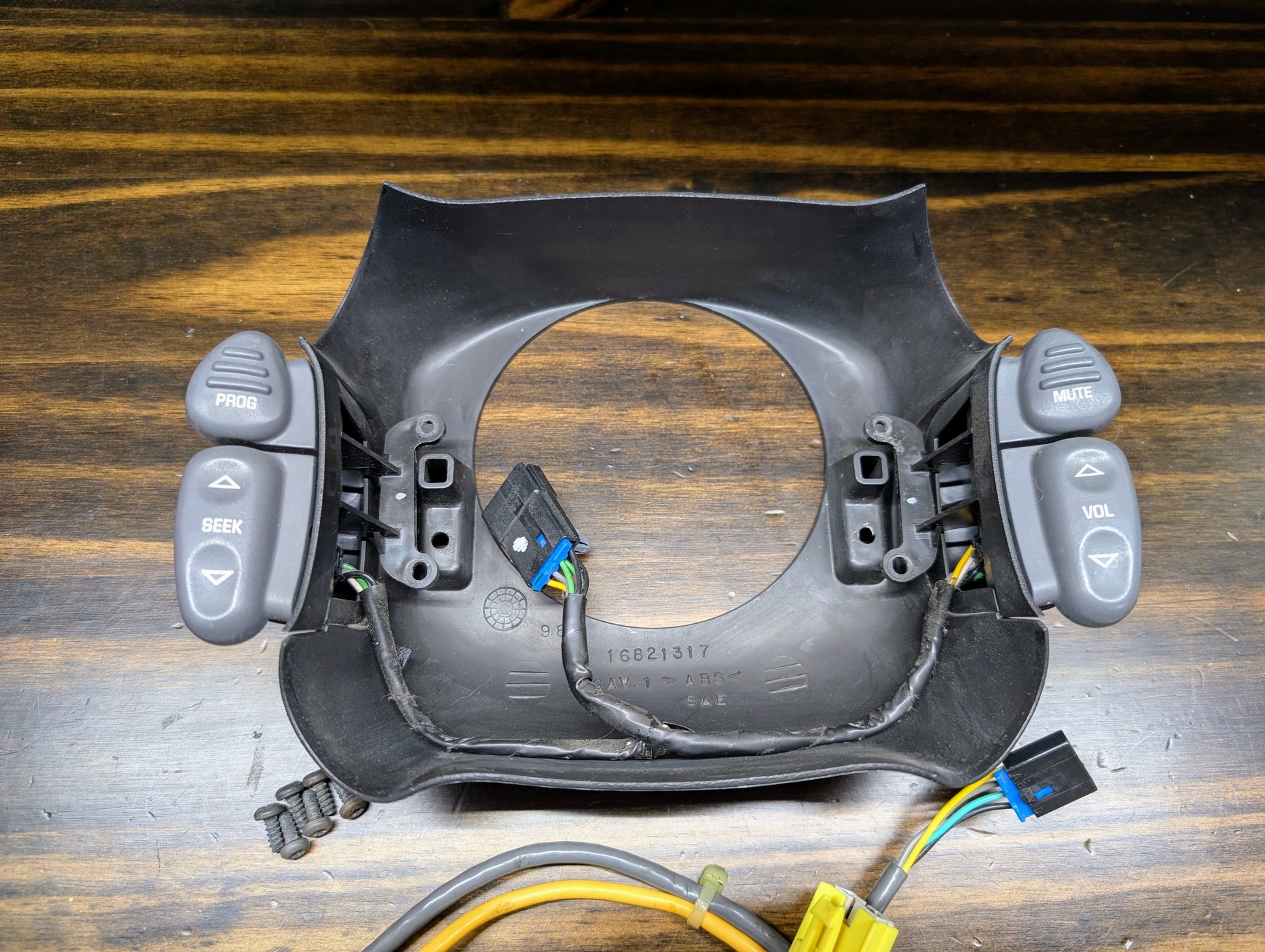A General Motors KIT Steering Wheel Radio Controls & Clock Spring for 1999 Chevy S10, Blazer, and more is shown on a wooden surface with exposed wires, connectors, four screws, and labeled buttons: PROG, SEEK, MUTE, VOL.