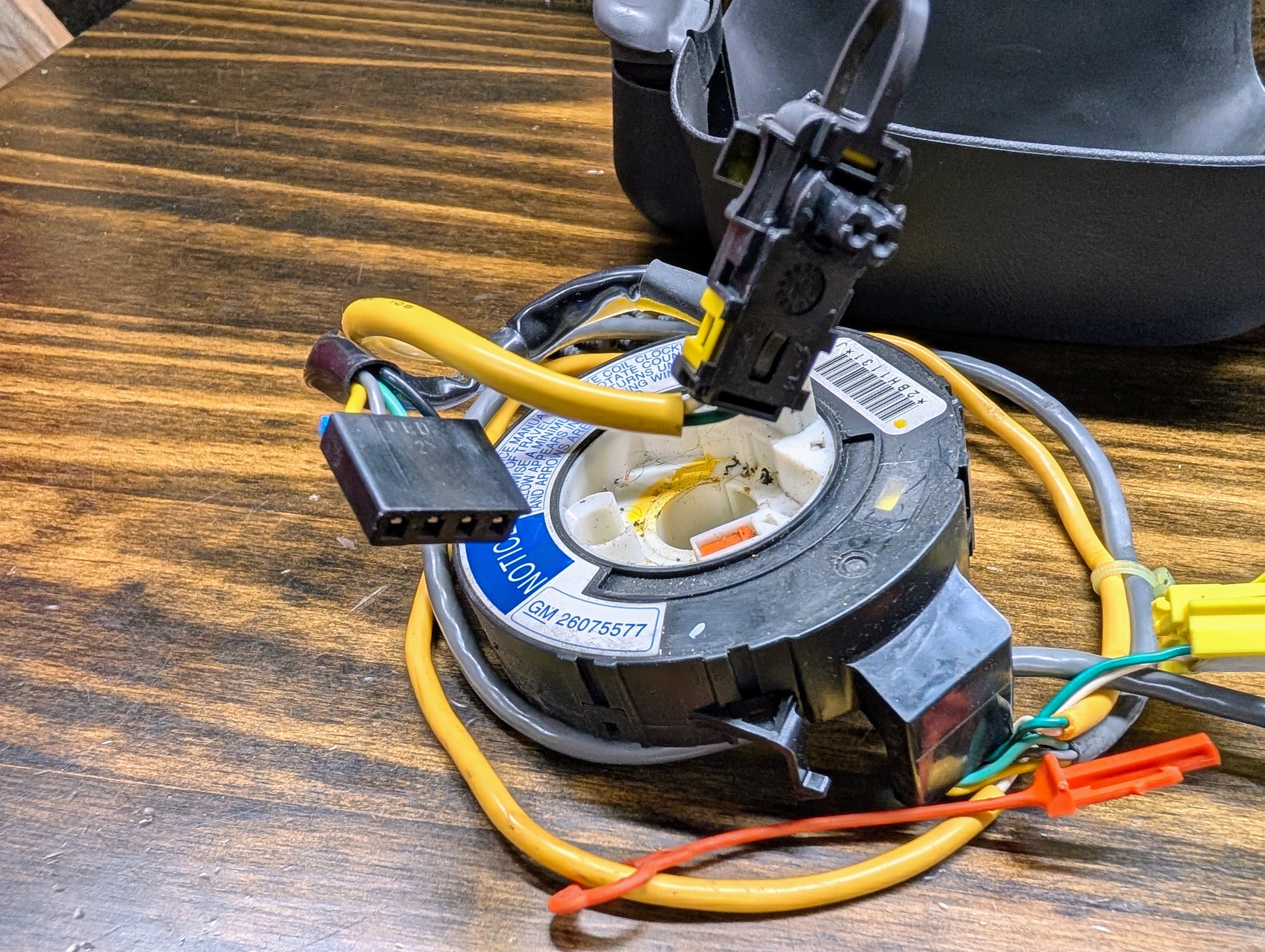 A close-up of the General Motors KIT Steering Wheel Radio Controls & Clock Spring for 1999 Chevy S10, Blazer, and more shows colored wires, connectors, and a barcode label on a wooden surface.